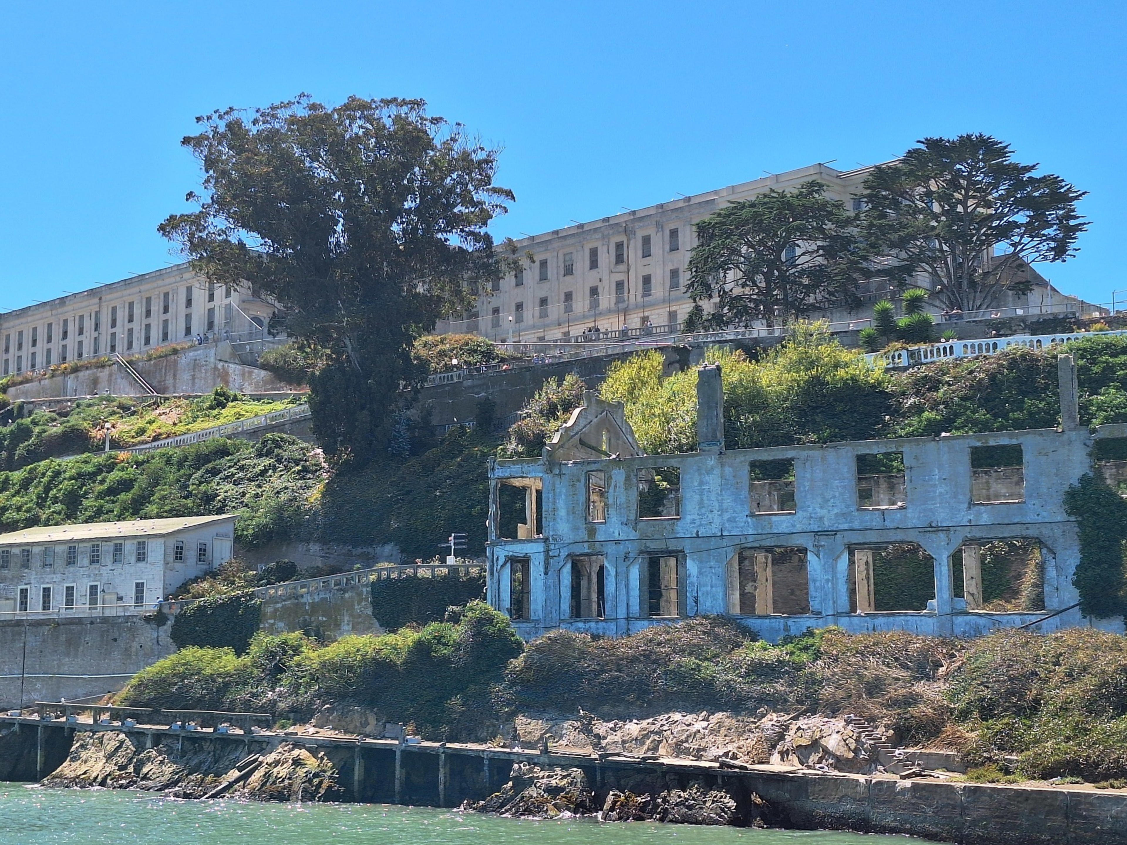 The image shows a large, old concrete building on a hill with overgrown greenery, partially surrounded by trees. Below it are the ruins of a smaller structure.