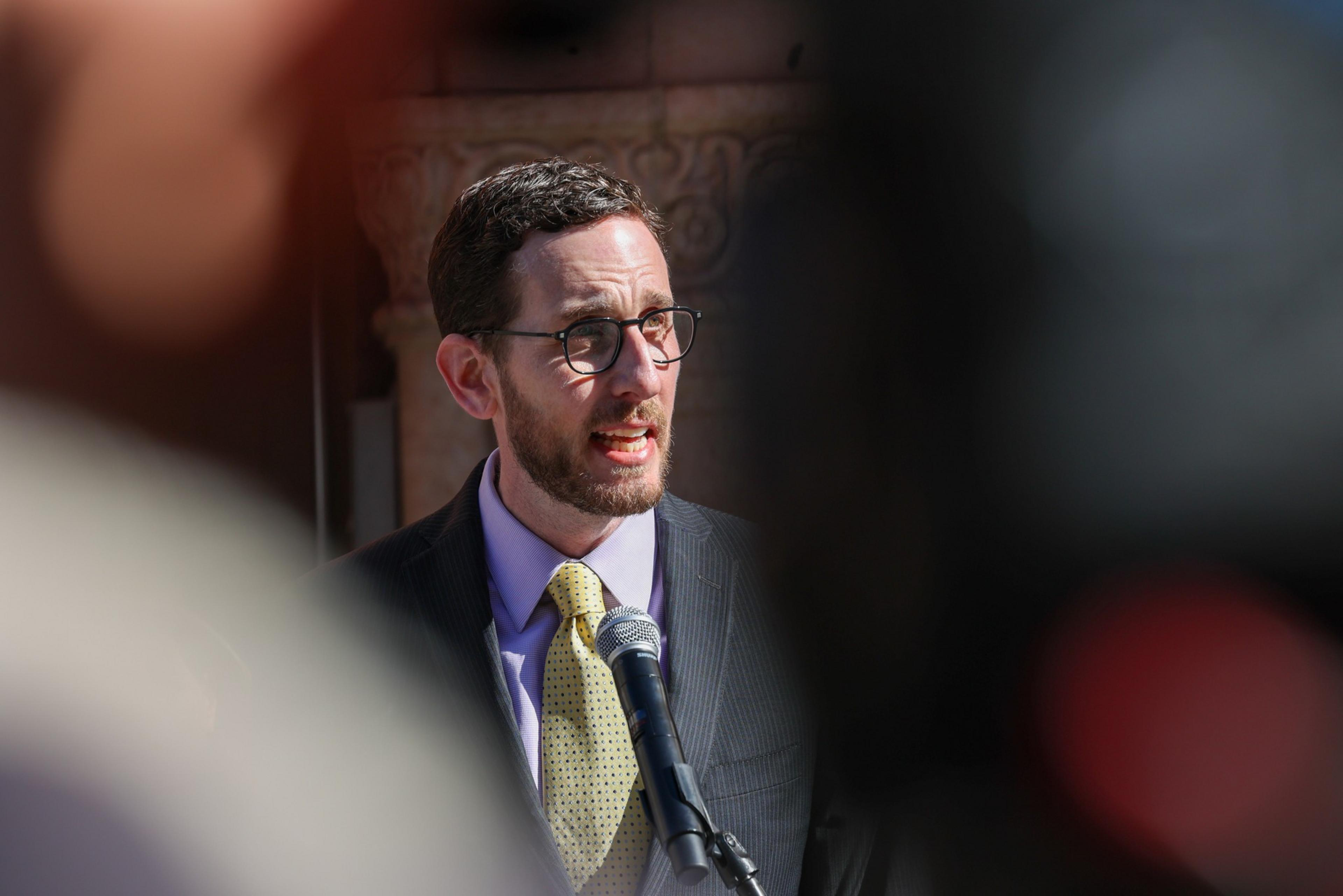 A man with glasses, a beard, and curly hair speaks into a microphone, wearing a suit and yellow tie, framed by blurred foreground shapes.