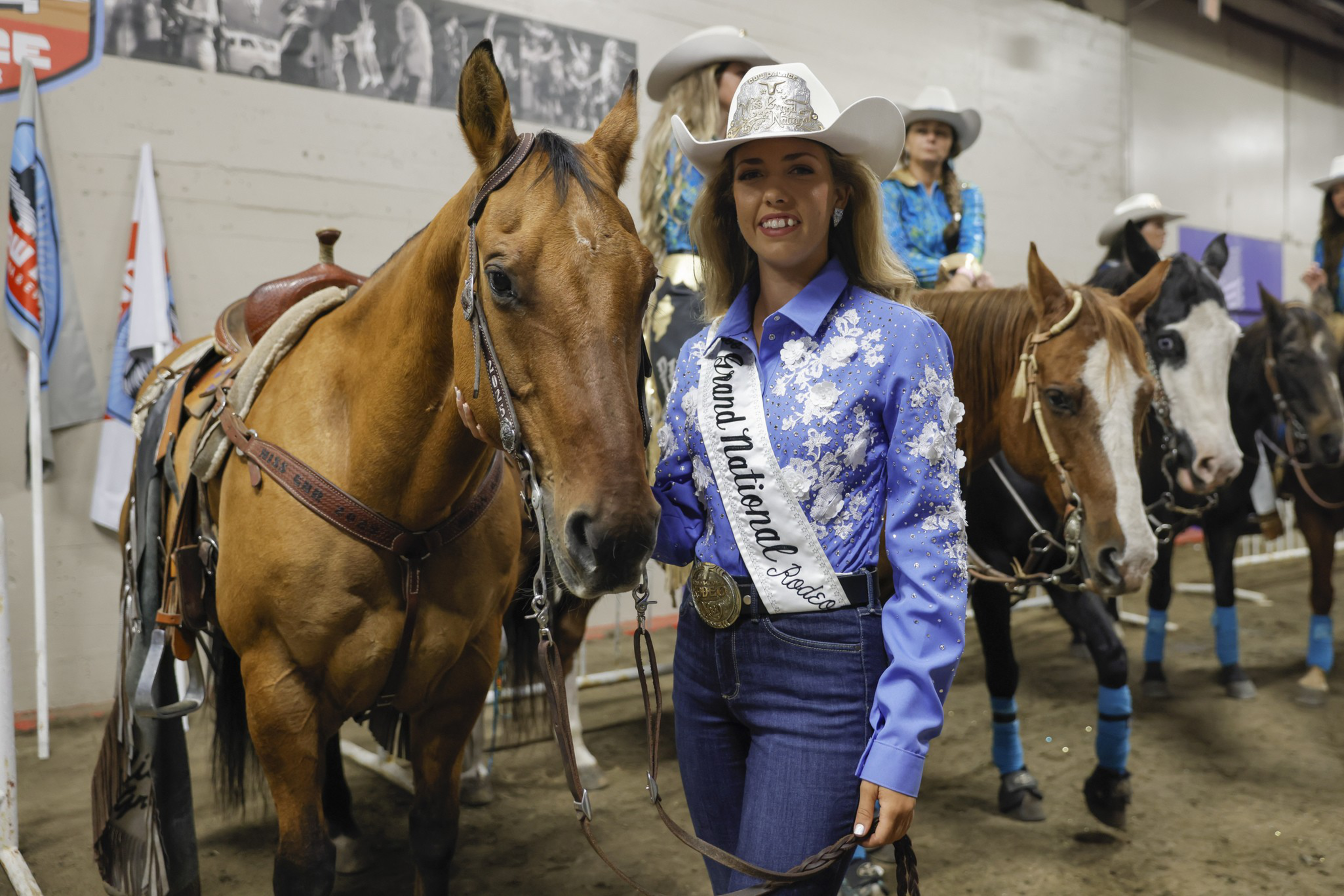 A young woman in a blue floral shirt, white cowboy hat, and a sash reading “Grand National Rodeo” stands beside a brown horse, with other riders and horses in the background.