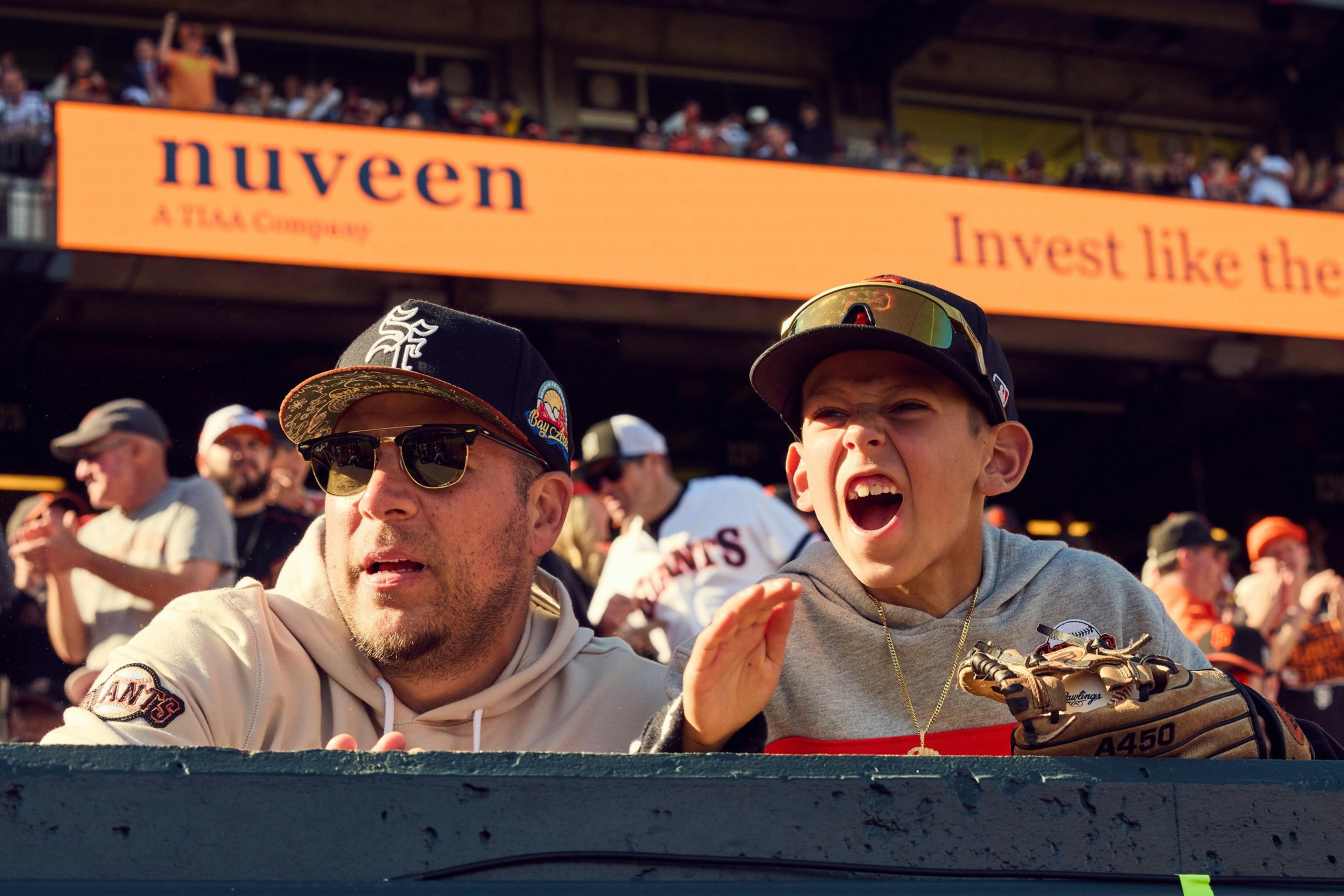 A man and a child at a sports game sit closely. The man wears sunglasses and a cap, while the child holds a baseball glove and appears excited.
