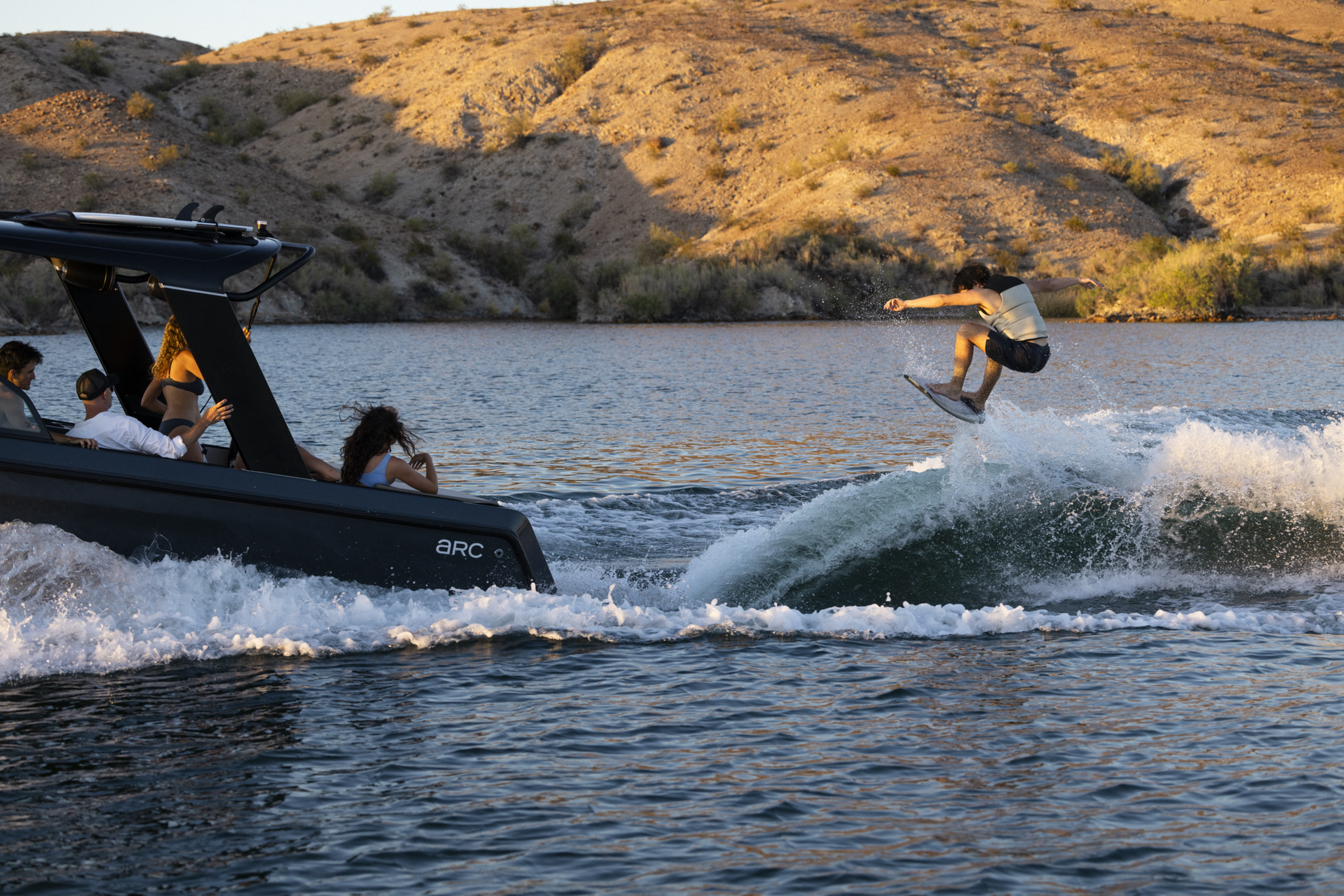 A person is wakesurfing behind a boat on a sunlit lake, surrounded by dry, hilly terrain. The boat has several passengers watching the surfer.