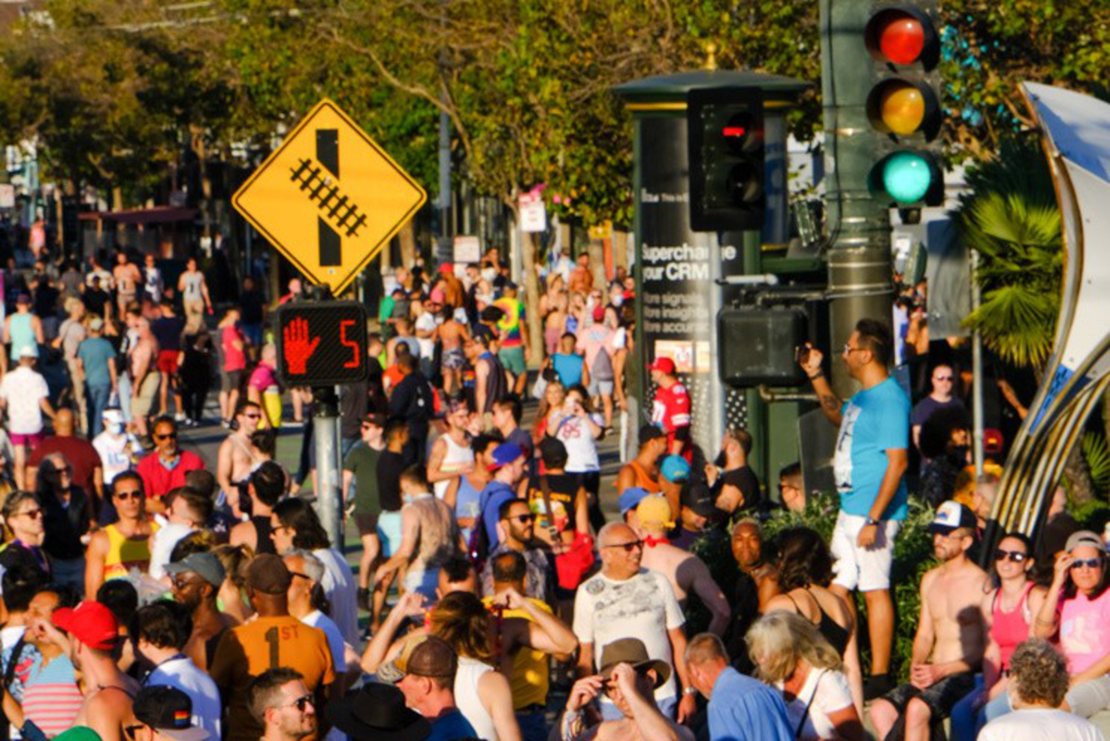 A large crowd of people stands tightly packed near a crossing with a red hand and countdown timer, beneath a traffic light showing green.