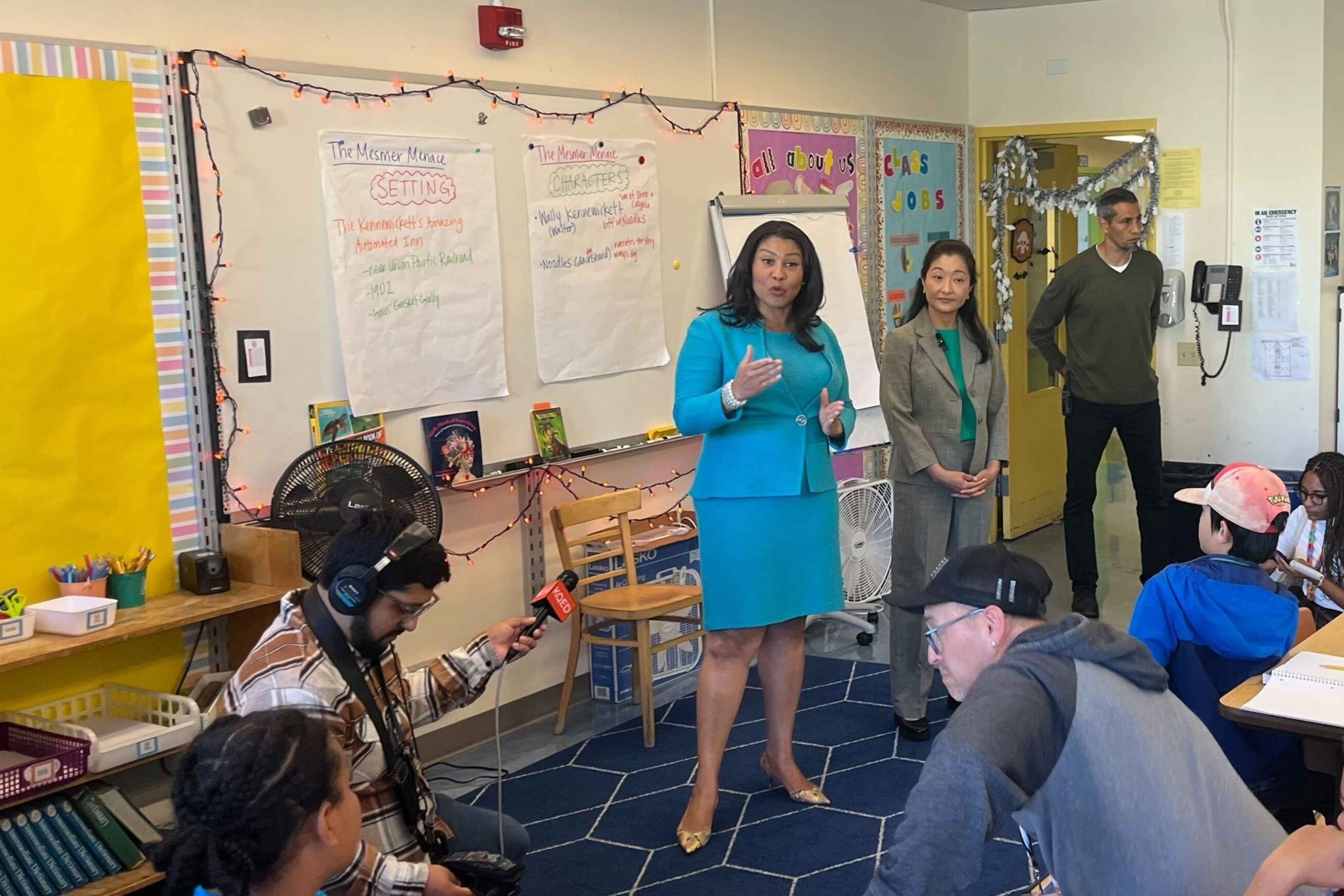 A woman in a blue outfit speaks to a classroom full of attentive students. A cameraman and another adult are present. The room is decorated with educational materials.