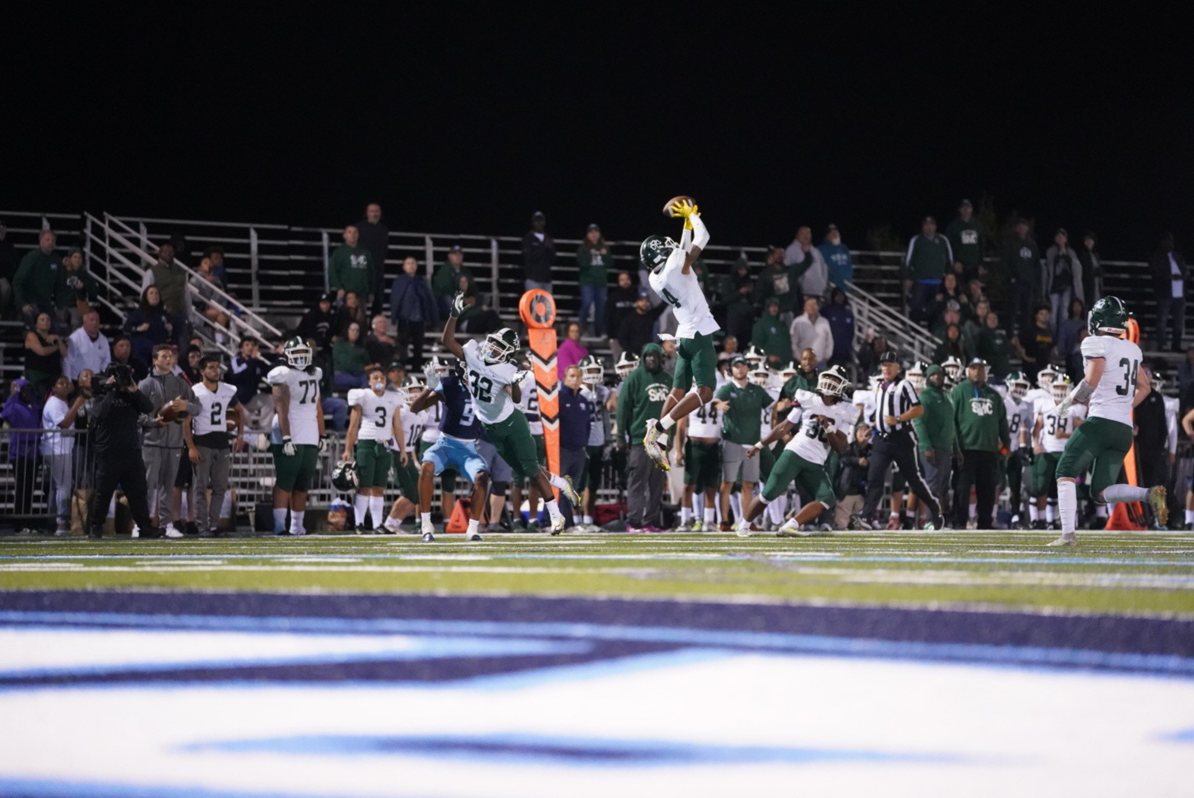 A football player in white and green jumps to catch the ball while defenders attempt to block him during a night game with spectators in the background.