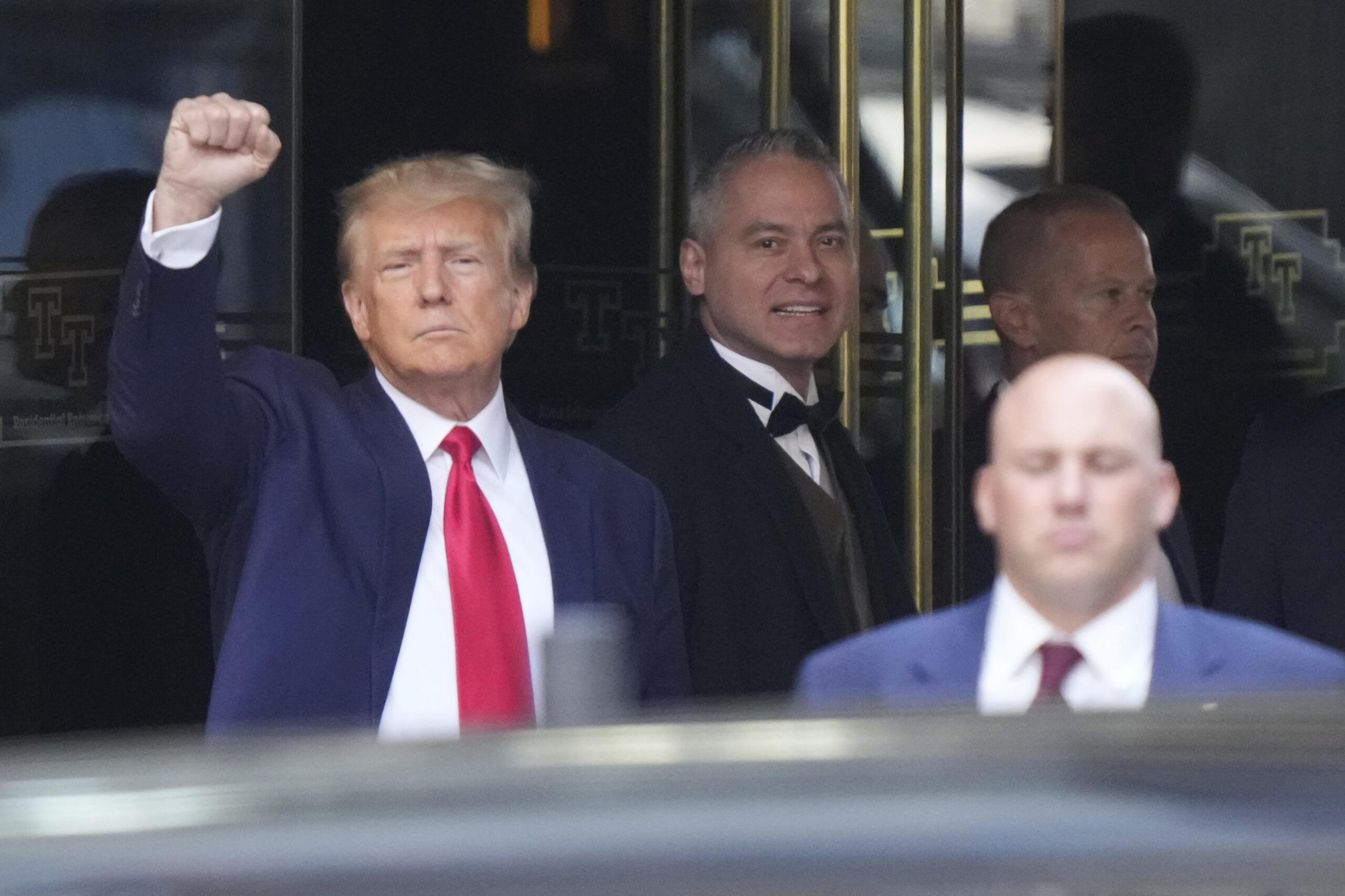 A man in a blue suit with a red tie raises his fist, while three other men in formal wear stand behind him near glass doors.