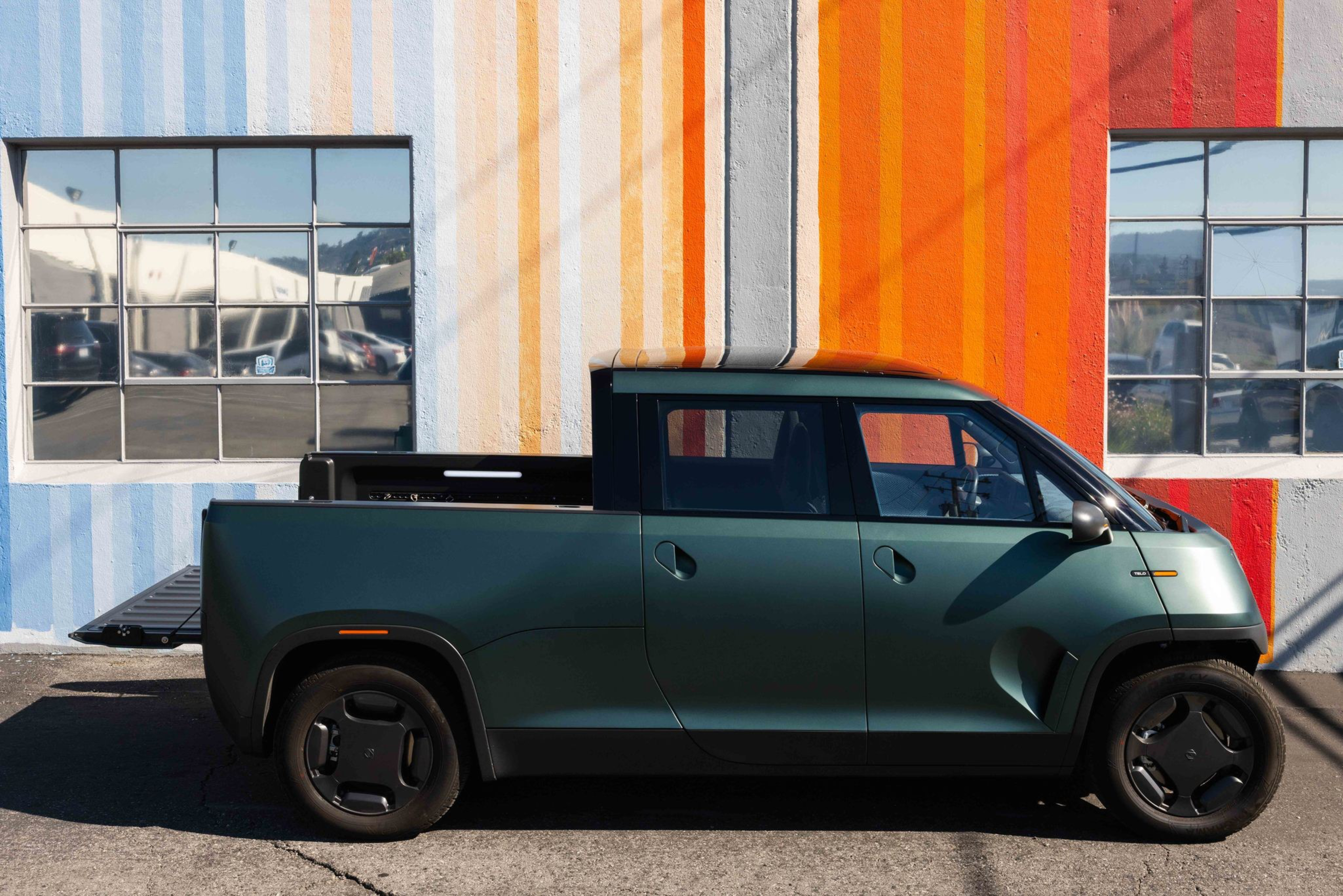 A dark green compact pickup truck is parked against a wall painted with vertical stripes in blue, yellow, gray, orange, and red, with two square windows.