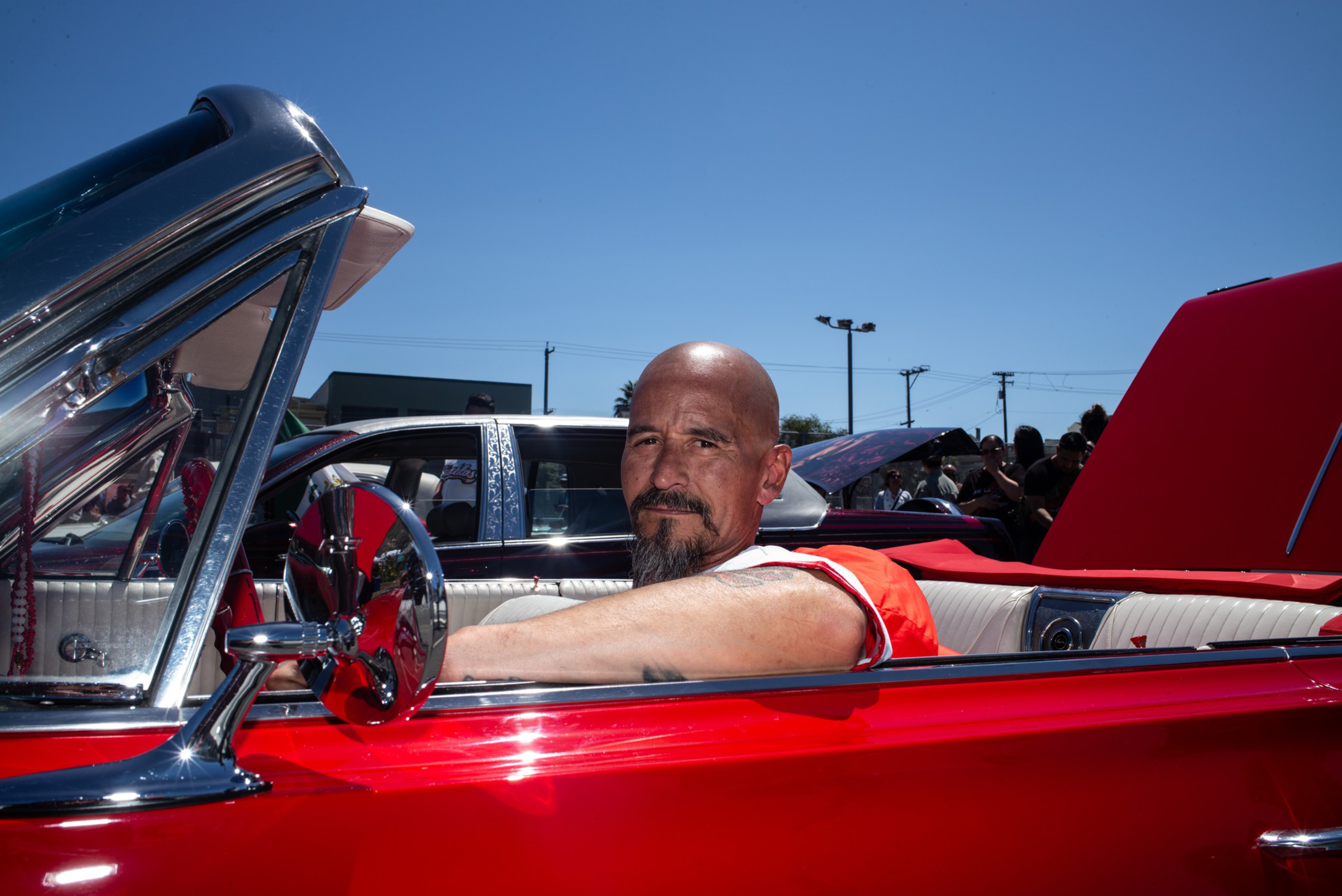 A bald man with a goatee sits in the driver’s seat of a shiny red convertible with the top down on a sunny day.
