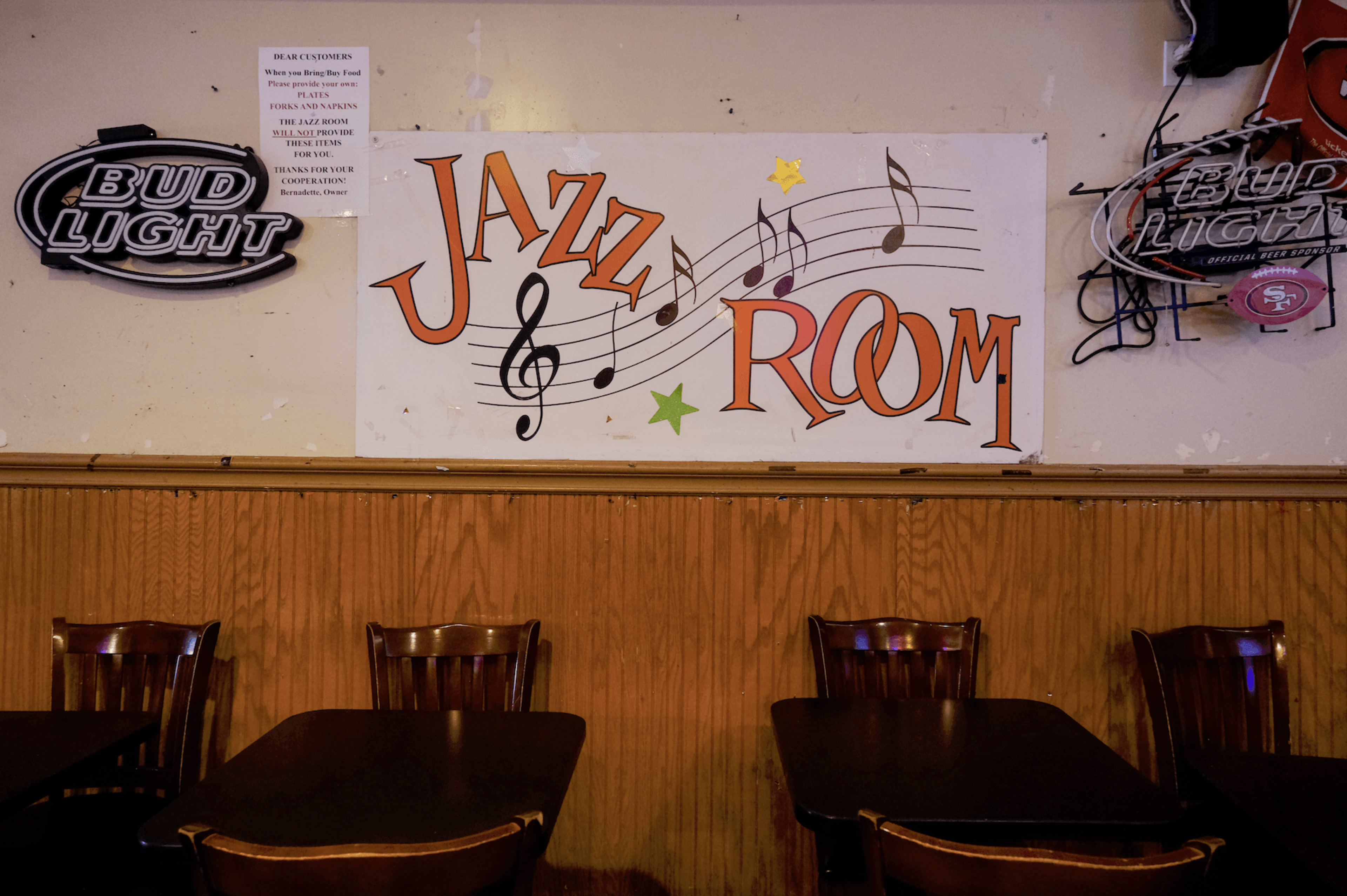 Four wooden chairs and dark tables are in front of a wall with a colorful "Jazz Room" sign and two Bud Light neon signs.
