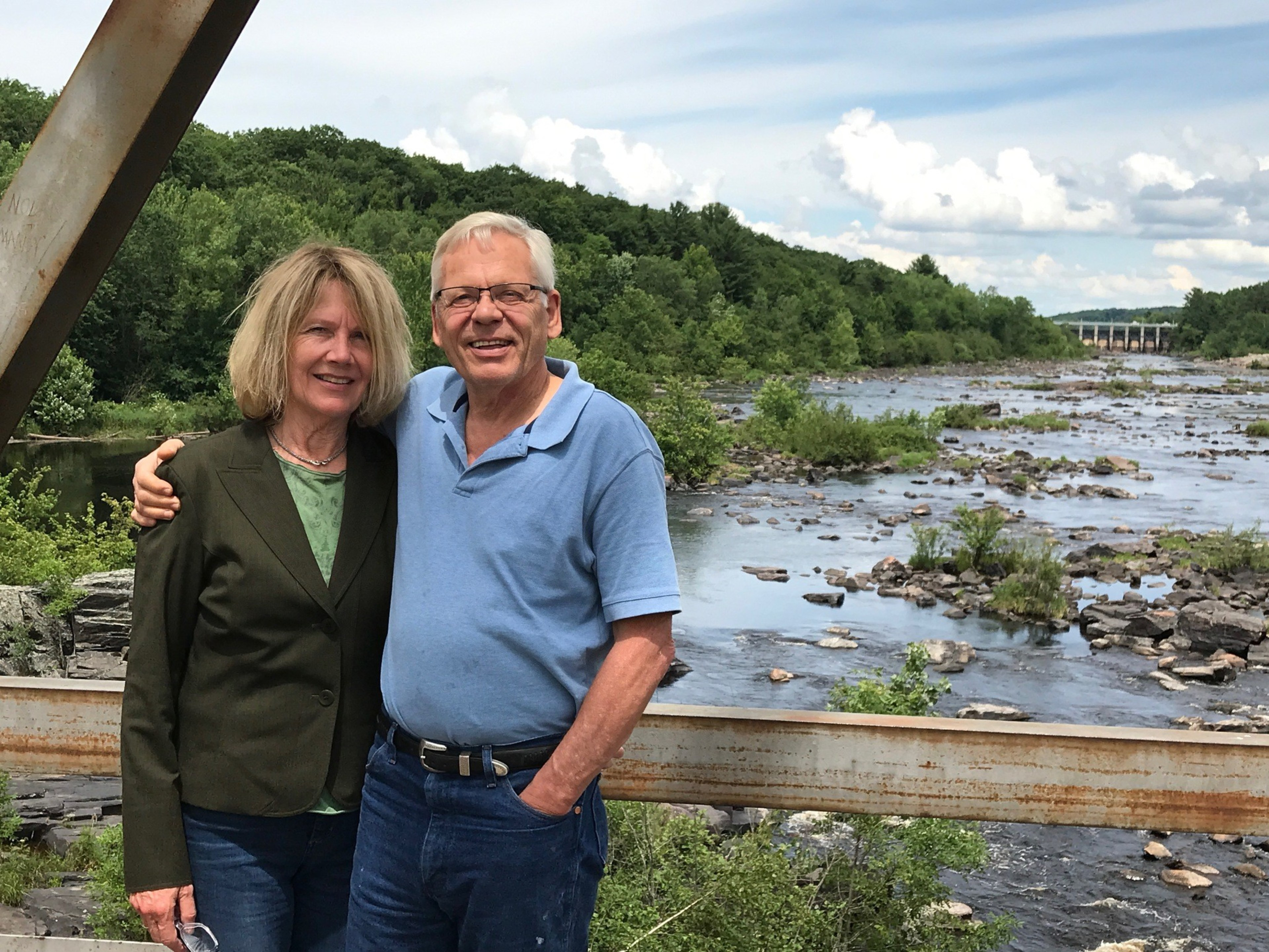 A couple poses happily near a river, surrounded by lush greenery and a cloudy sky. One wears a dark jacket, the other a light blue shirt.