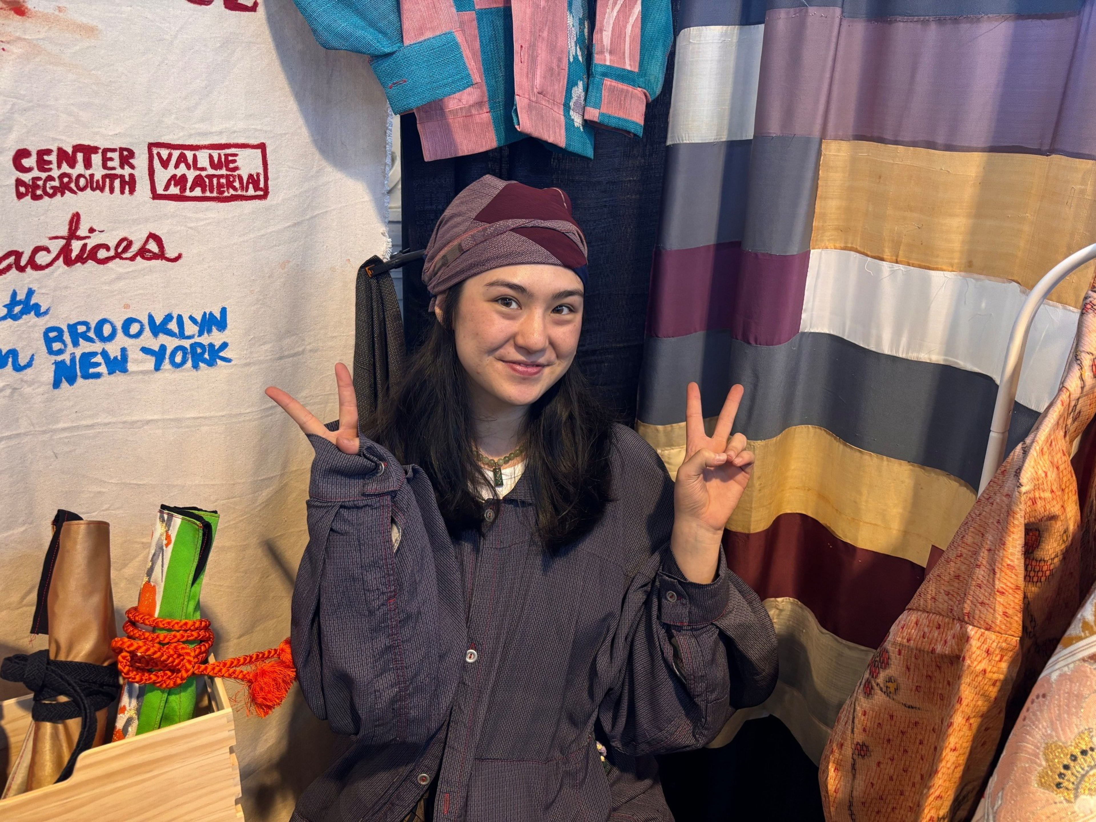 A smiling woman wearing a headscarf and loose dark shirt flashes peace signs with both hands in front of colorful fabric and wall art.