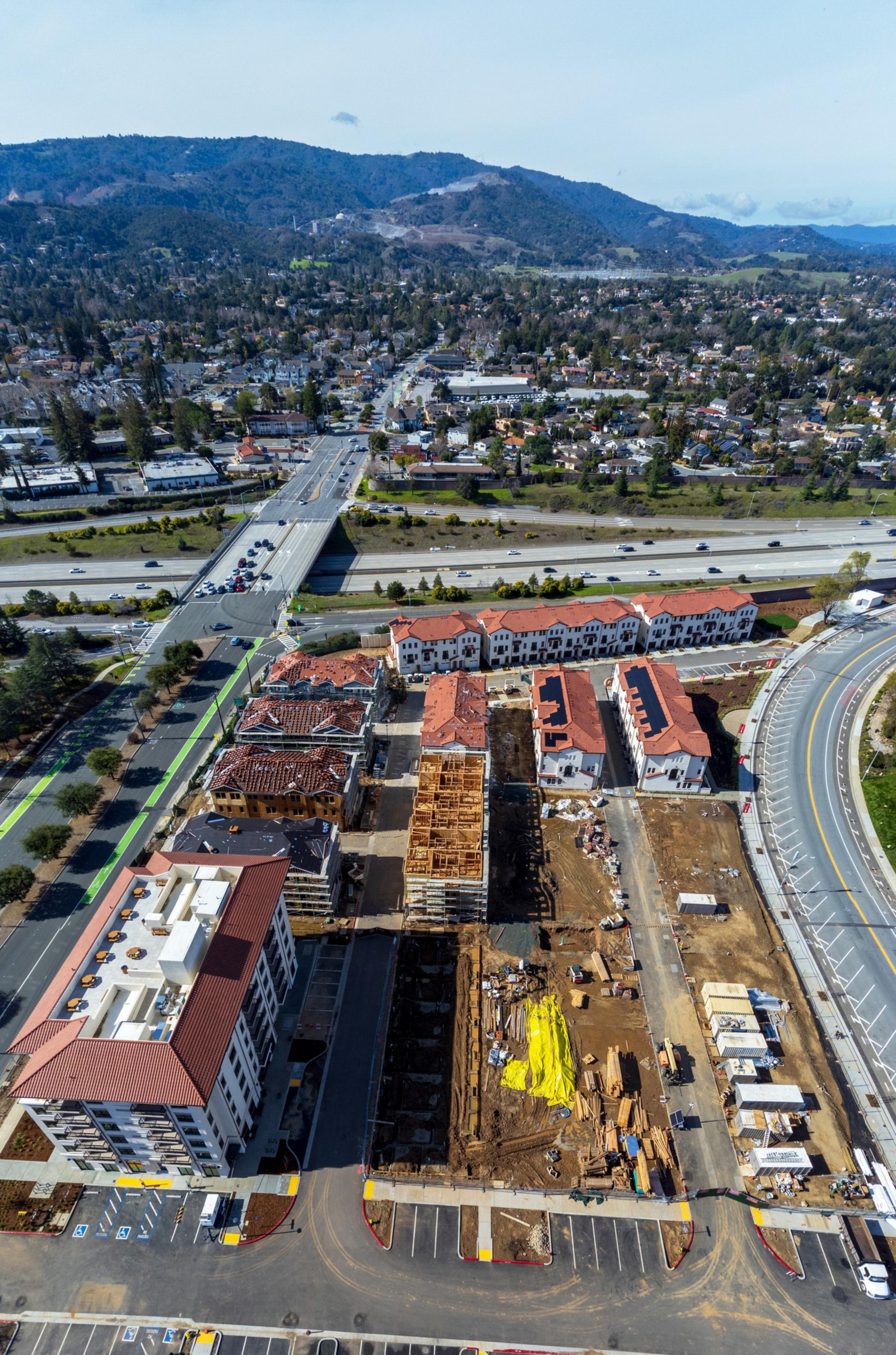 The image shows an aerial view of a suburban area with construction sites, roads with vehicles, and residential buildings. Hills are visible in the distance.
