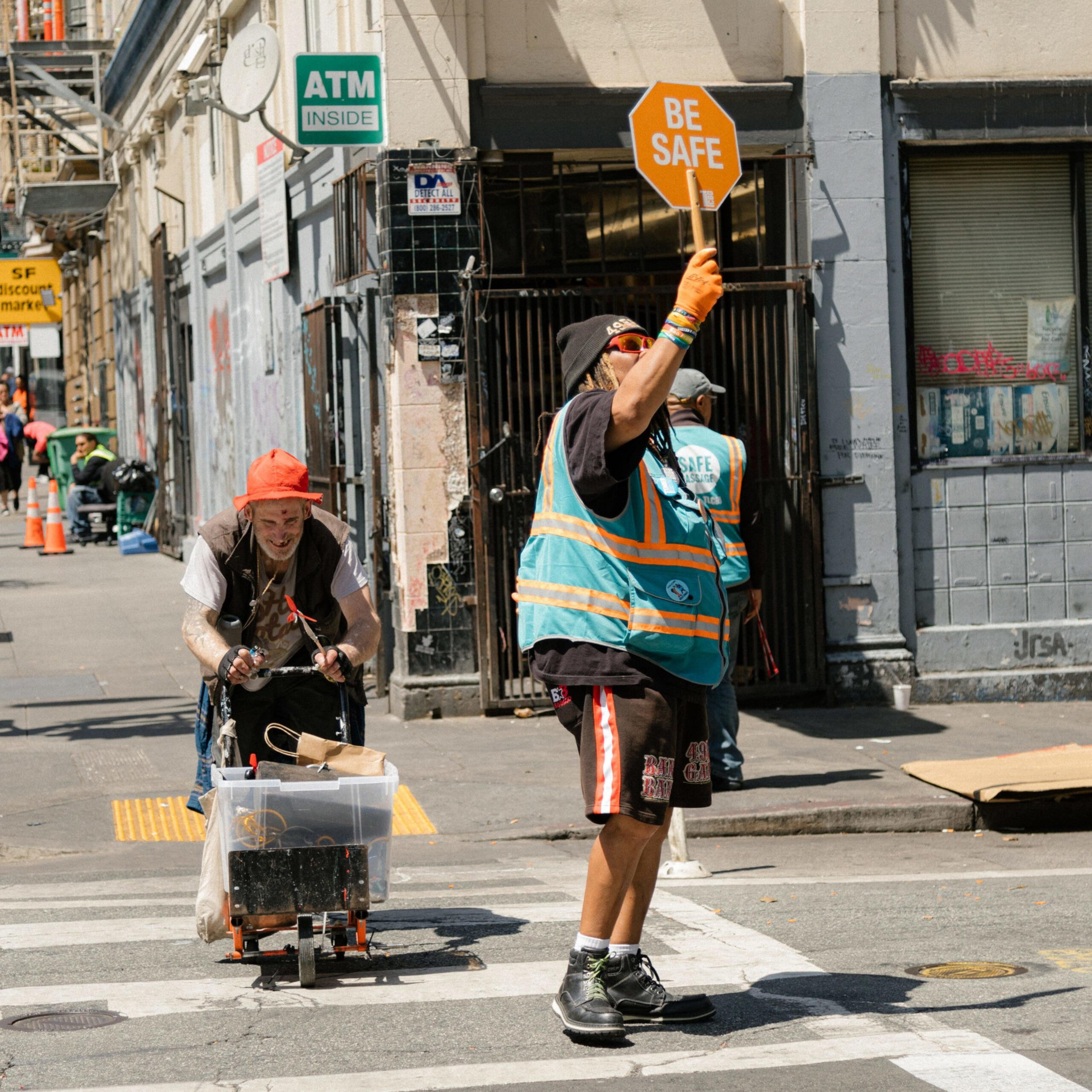 A person in a safety vest holds a "BE SAFE" sign on a crosswalk while another person with an orange hat pushes a loaded cart behind them.