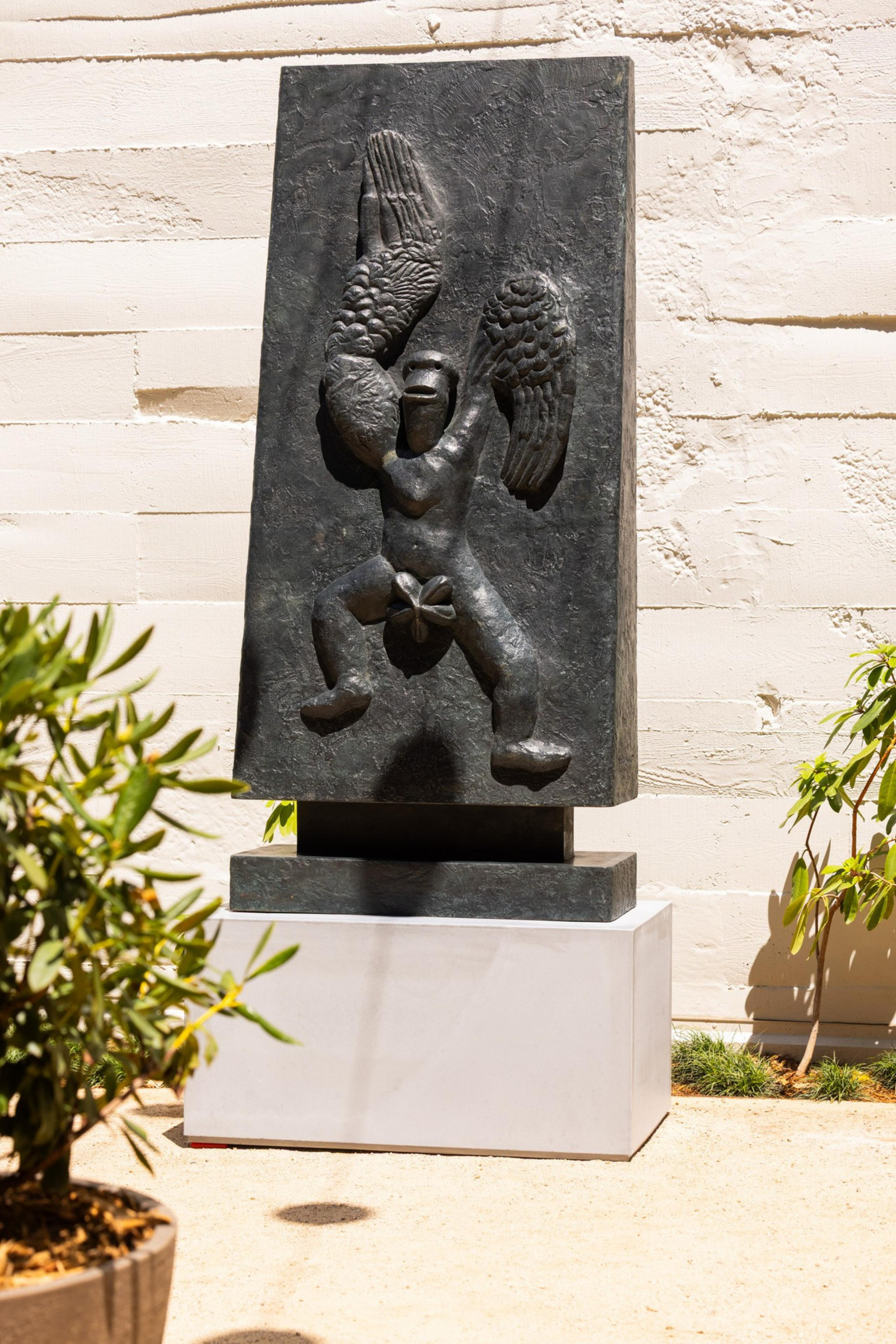 The image shows a dark sculpture of a human figure with wings, carved into a rectangular slab. It's outdoors, with potted plants nearby.