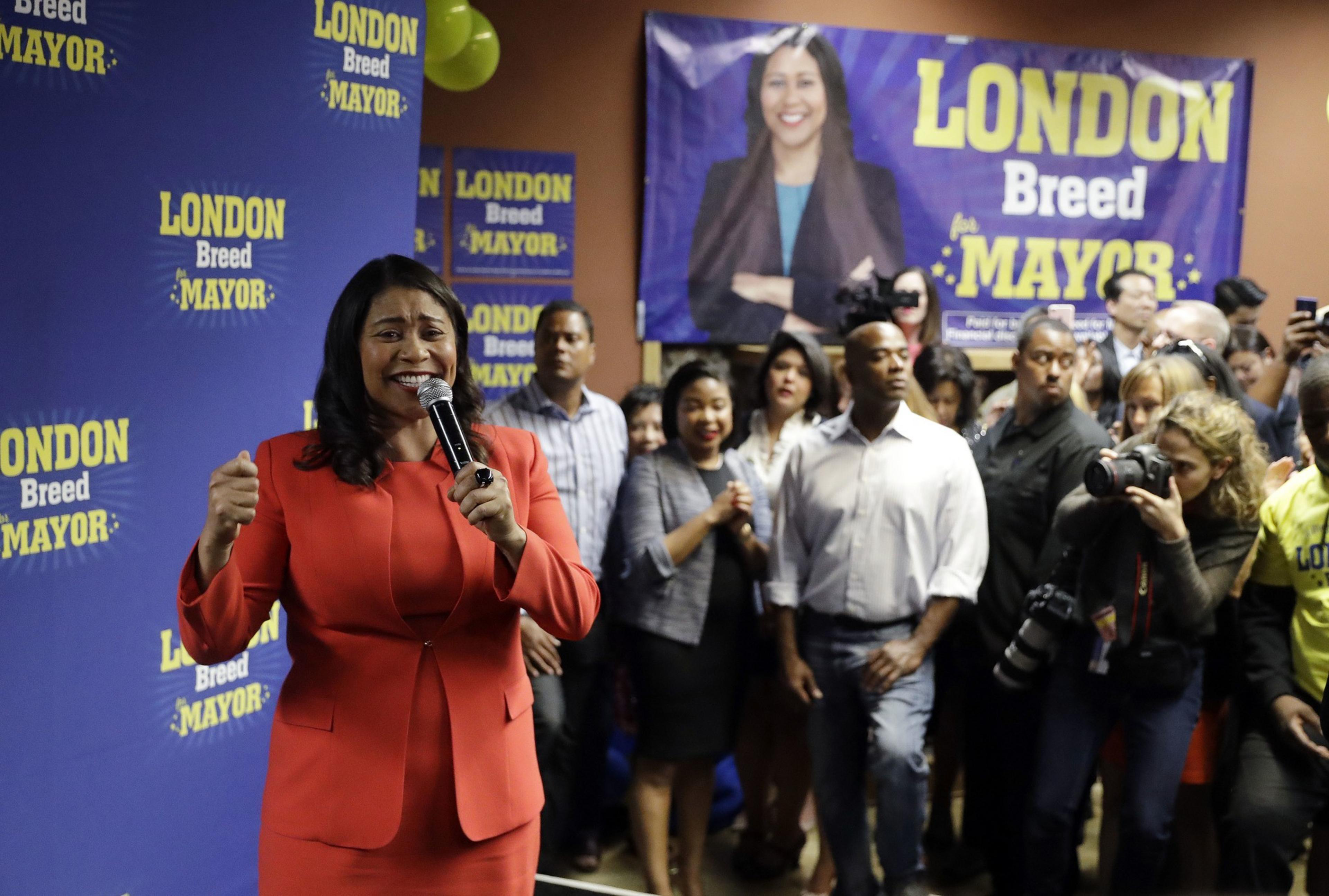 A woman in a red outfit is speaking into a microphone at a campaign event with a supportive crowd and a large poster behind her.
