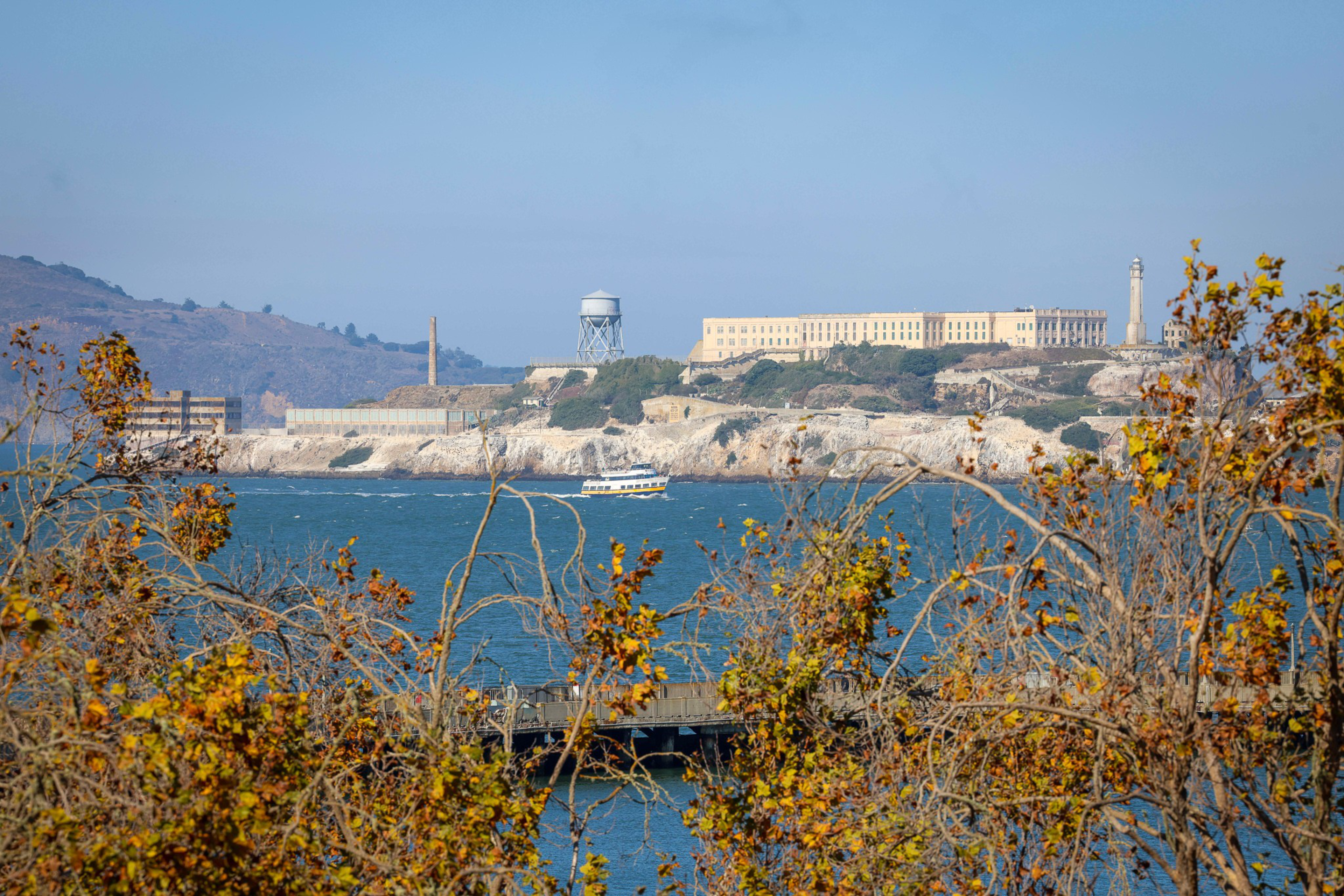 A large historic prison building sits on an island across blue water, with a ferry boat nearby and trees with autumn leaves framing the view.