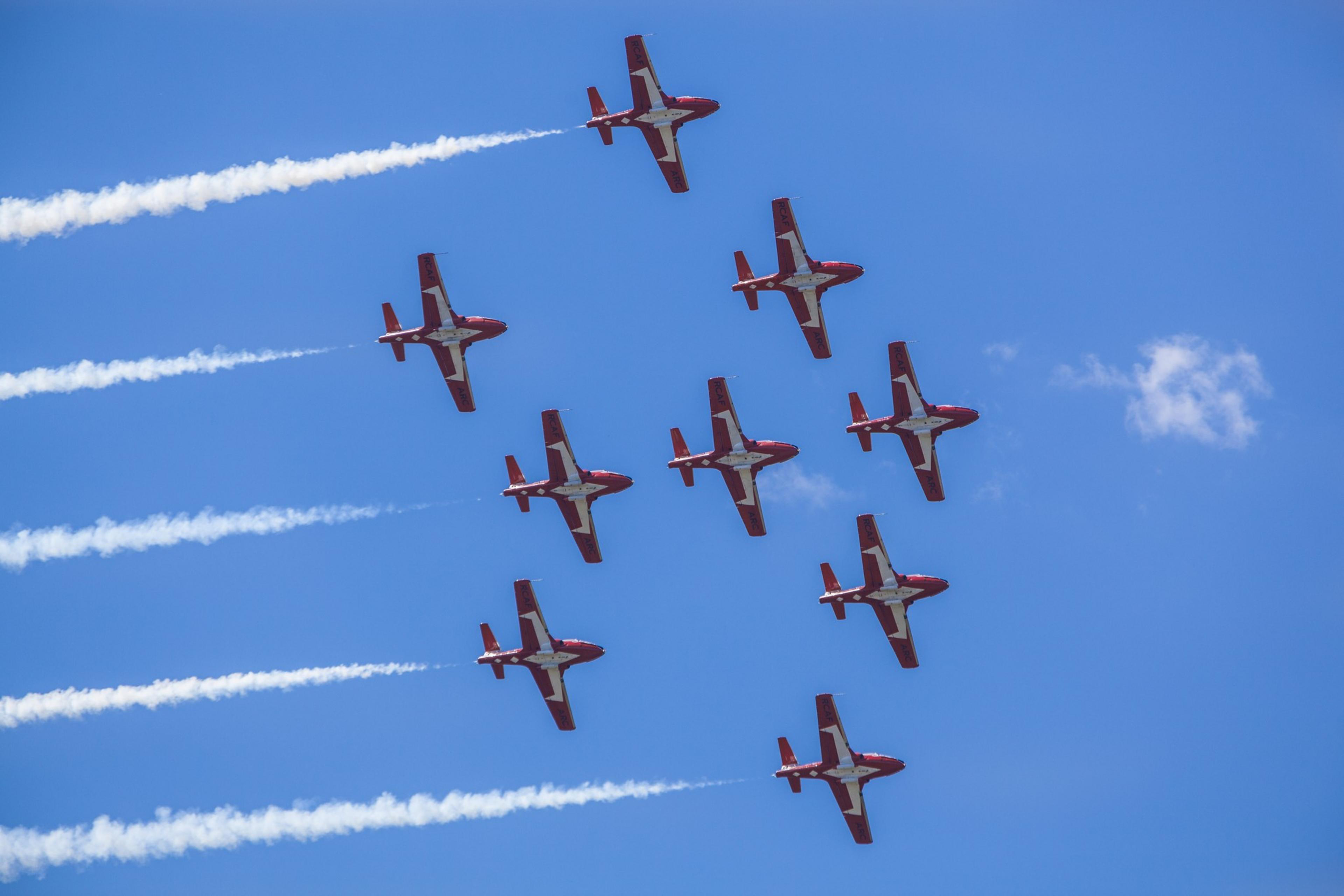 Nine red and white airplanes fly in close formation against a clear blue sky, leaving white smoke trails behind them.