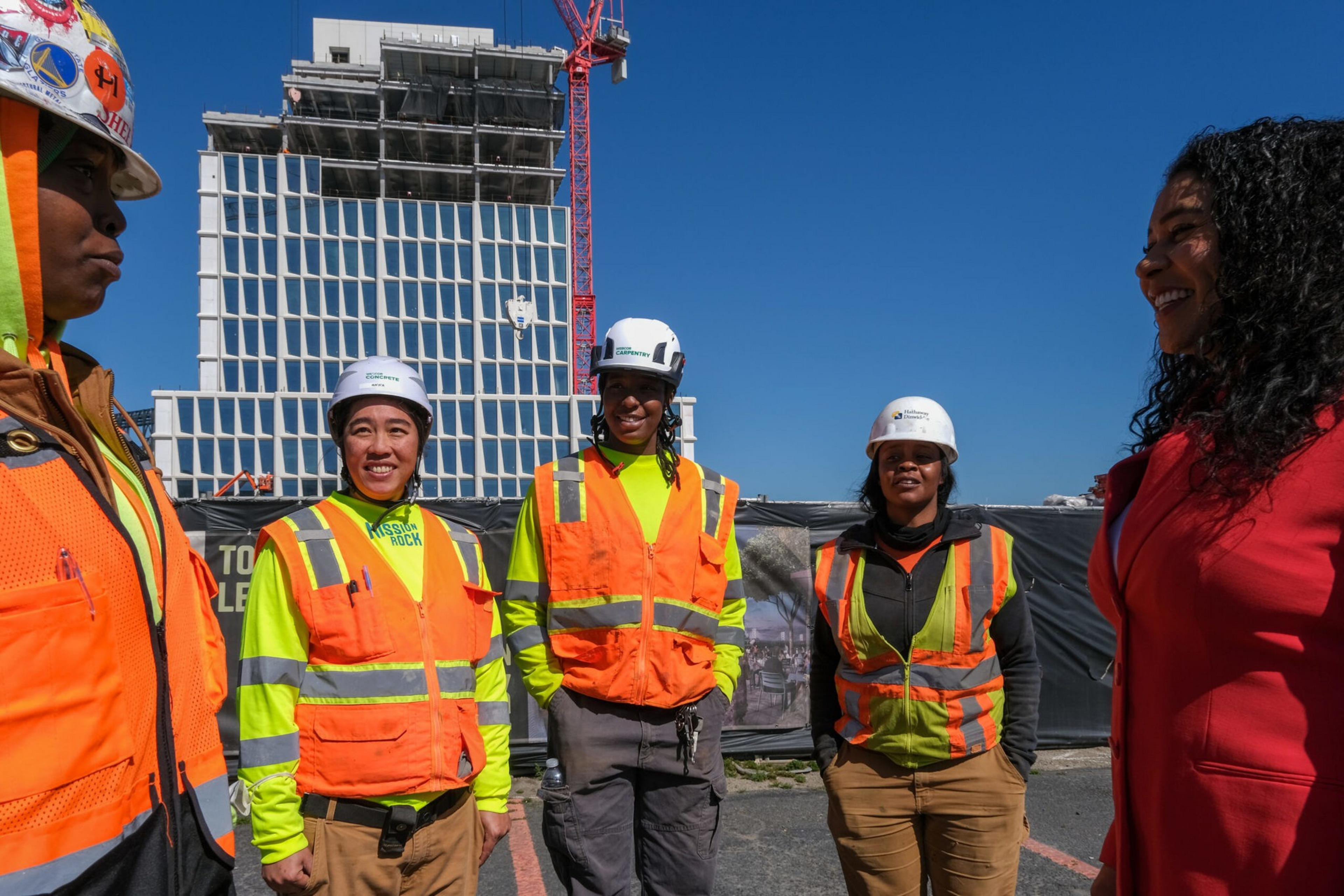 Five people, four in neon yellow and orange construction gear and one in a red jacket, stand smiling outdoors by a construction site.