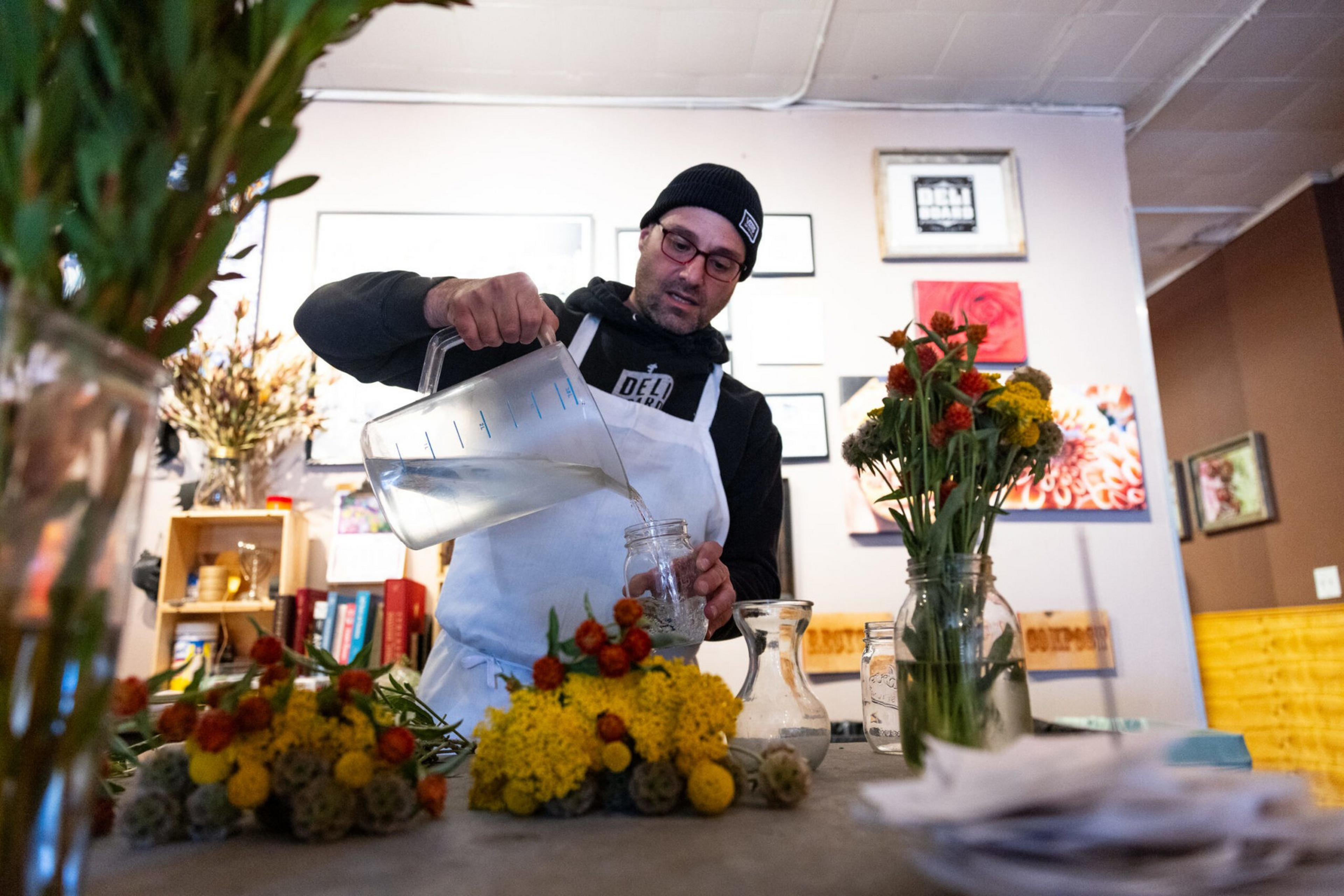 A man in a black beanie and white apron pours water from a pitcher into a jar, surrounded by colorful flower arrangements on a table.