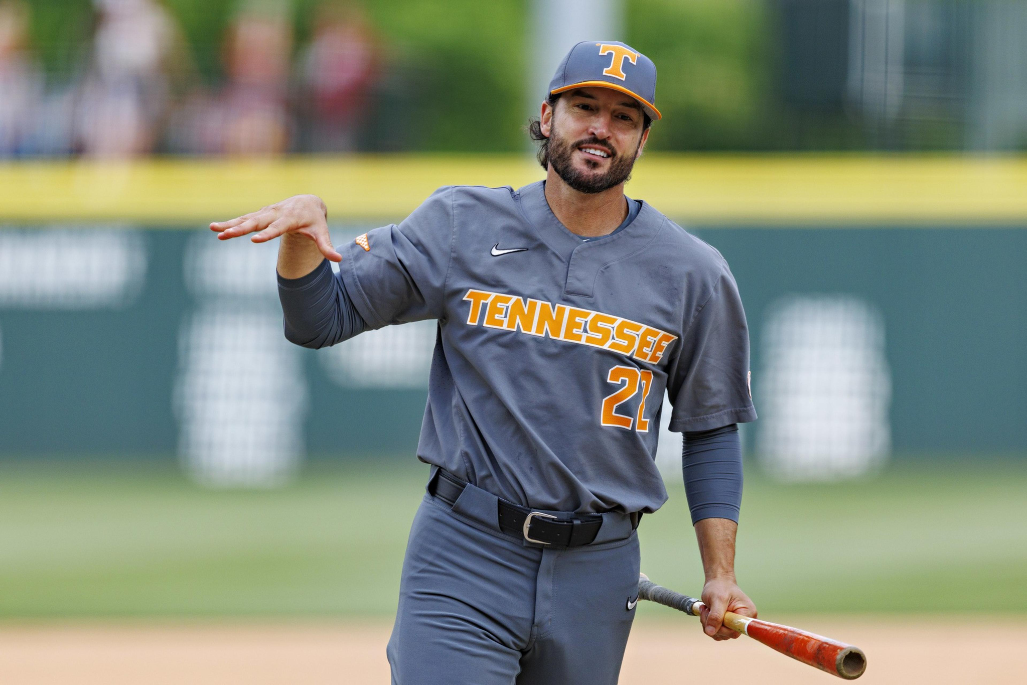 A baseball player wearing a gray "Tennessee" uniform with number 21 holds a bat and gestures with his left hand on the field.