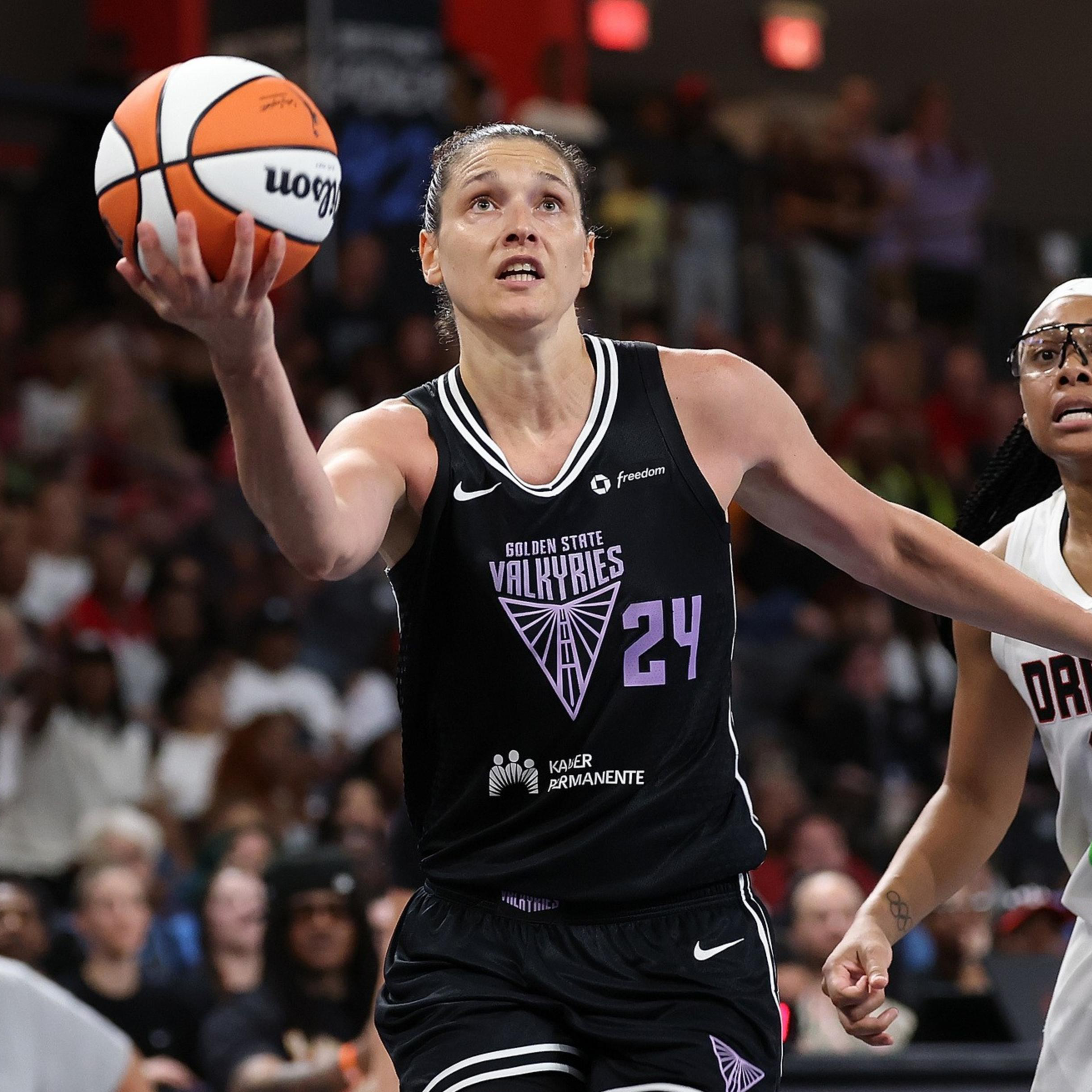 A player in a black Golden State Valkyries jersey leaps with a basketball toward the basket, while a player in a white Dream jersey closely defends her.