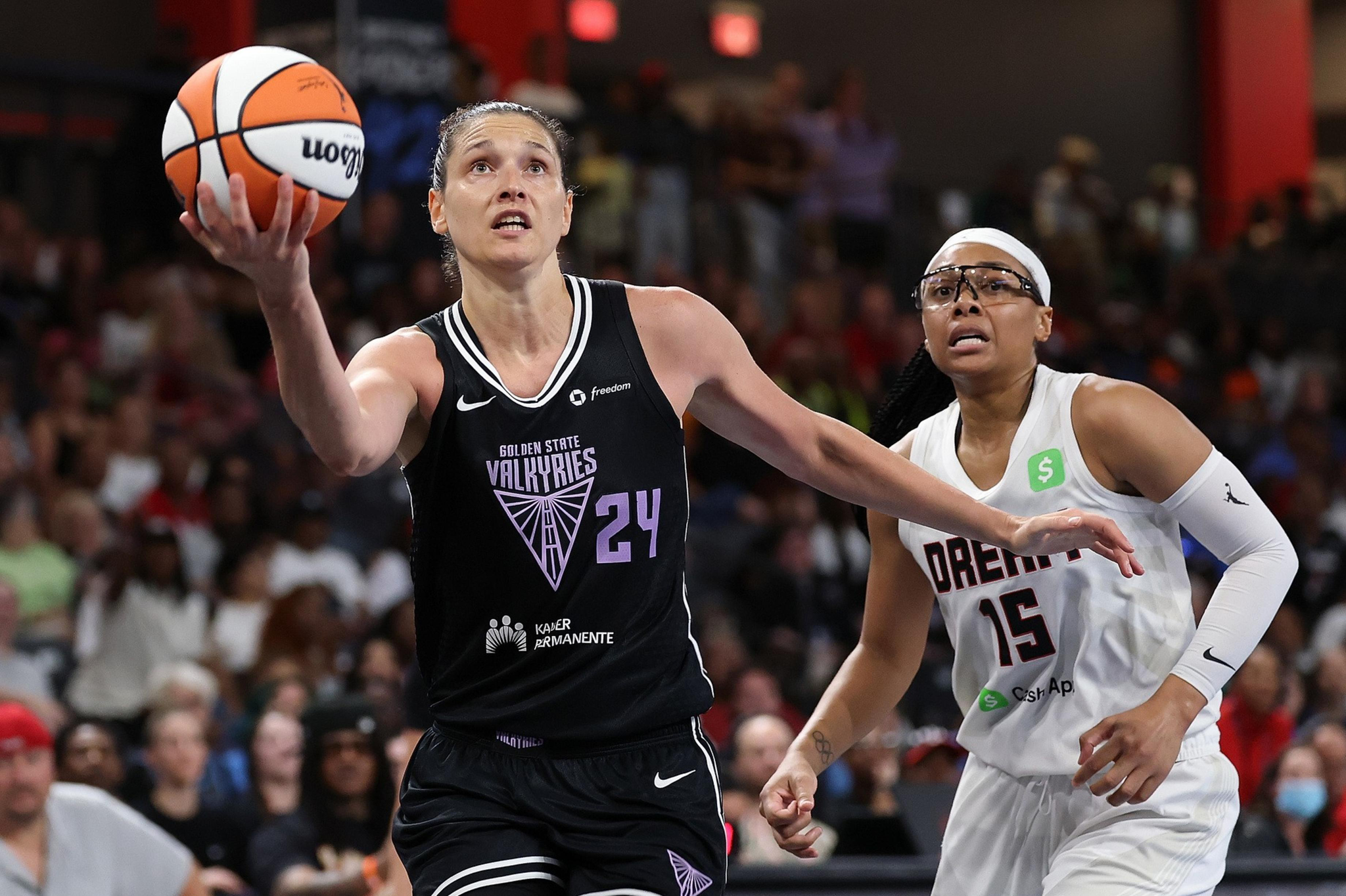 A player in a black Golden State Valkyries jersey leaps with a basketball toward the basket, while a player in a white Dream jersey closely defends her.