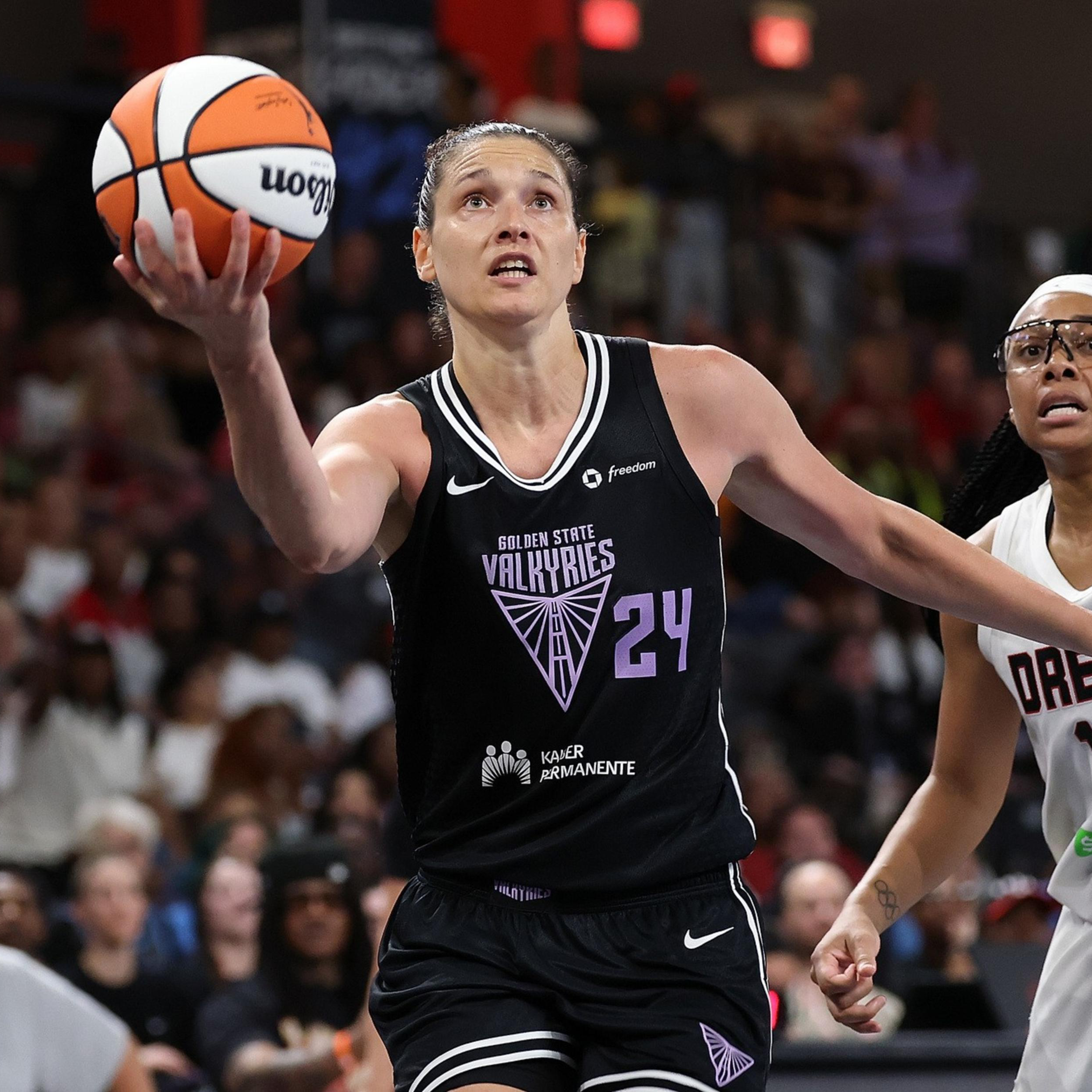 A player in a black Golden State Valkyries jersey leaps with a basketball toward the basket, while a player in a white Dream jersey closely defends her.