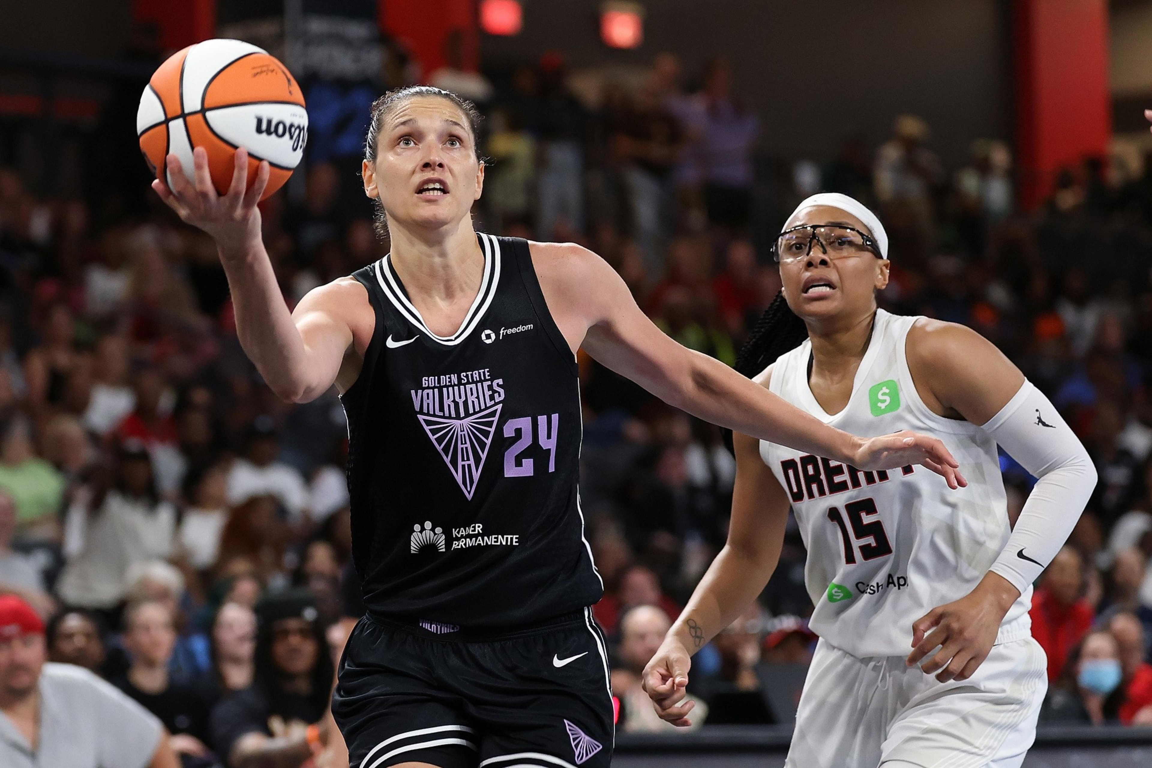 A player in a black Golden State Valkyries jersey leaps with a basketball toward the basket, while a player in a white Dream jersey closely defends her.