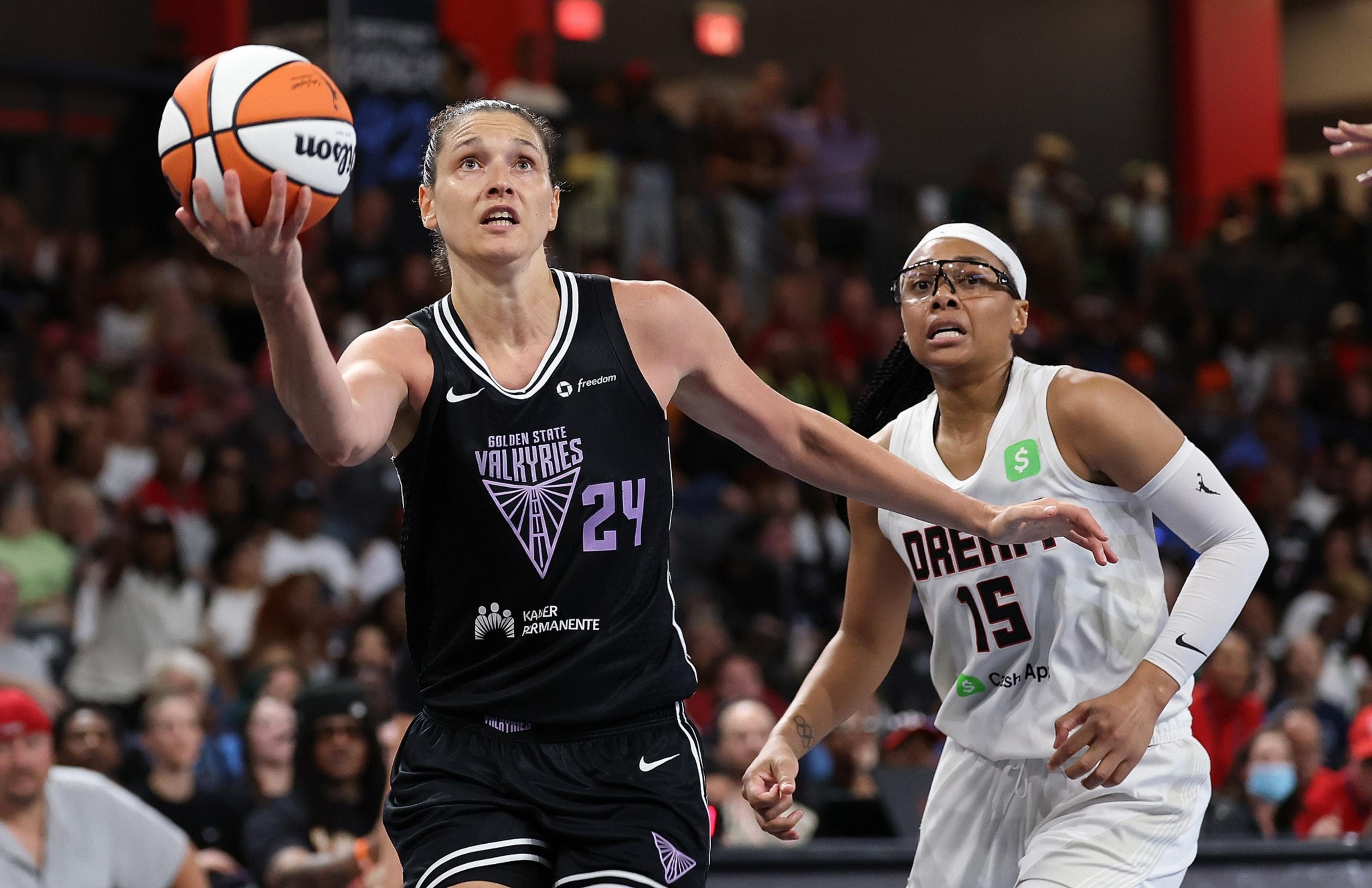 A player in a black Golden State Valkyries jersey leaps with a basketball toward the basket, while a player in a white Dream jersey closely defends her.