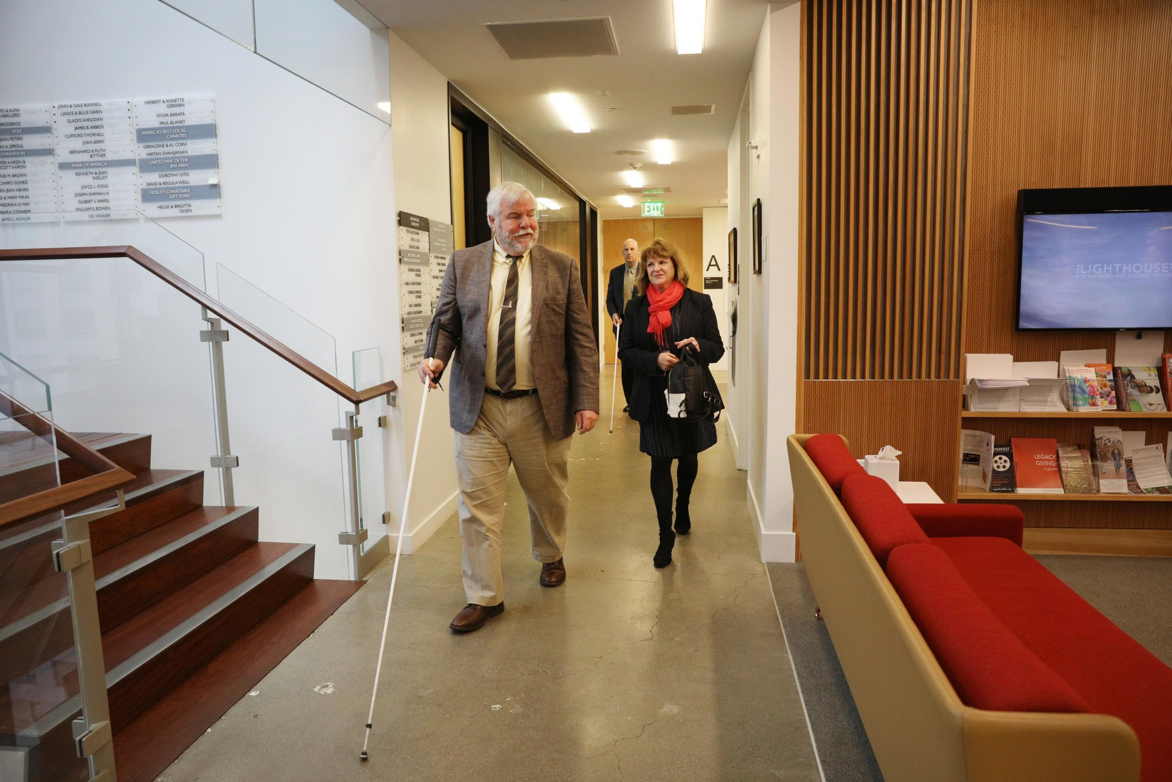A man with a white cane walks through a modern hallway with a woman. They're casually dressed, and there are stairs, signage, and a display with brochures nearby.