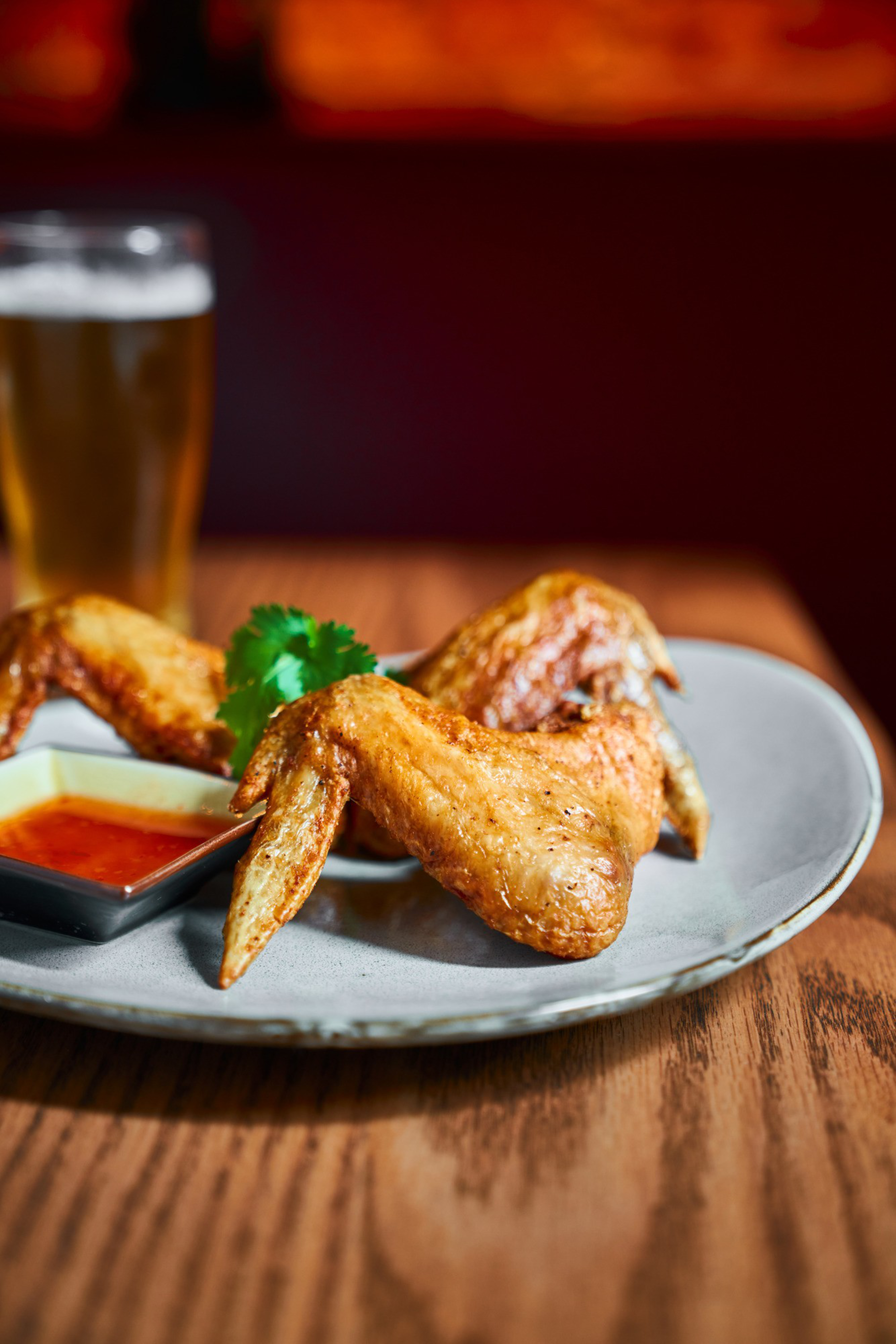 Crispy chicken wings with a sprig of cilantro sit on a plate, accompanied by a small dish of dipping sauce. A glass of beer is visible in the background.