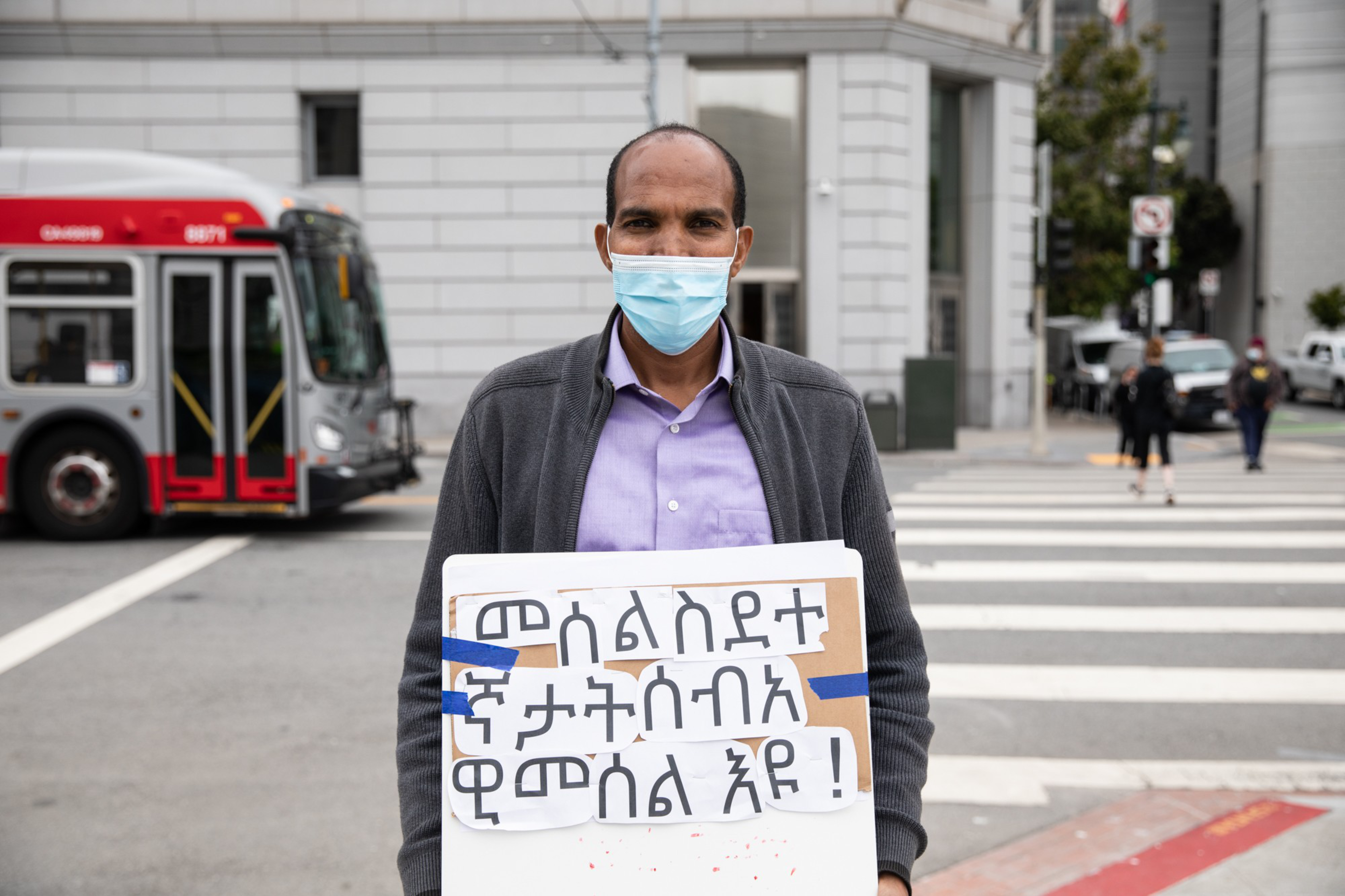 A man wearing a face mask holds a sign with Amharic text while standing on a city street near a crosswalk and a red bus.