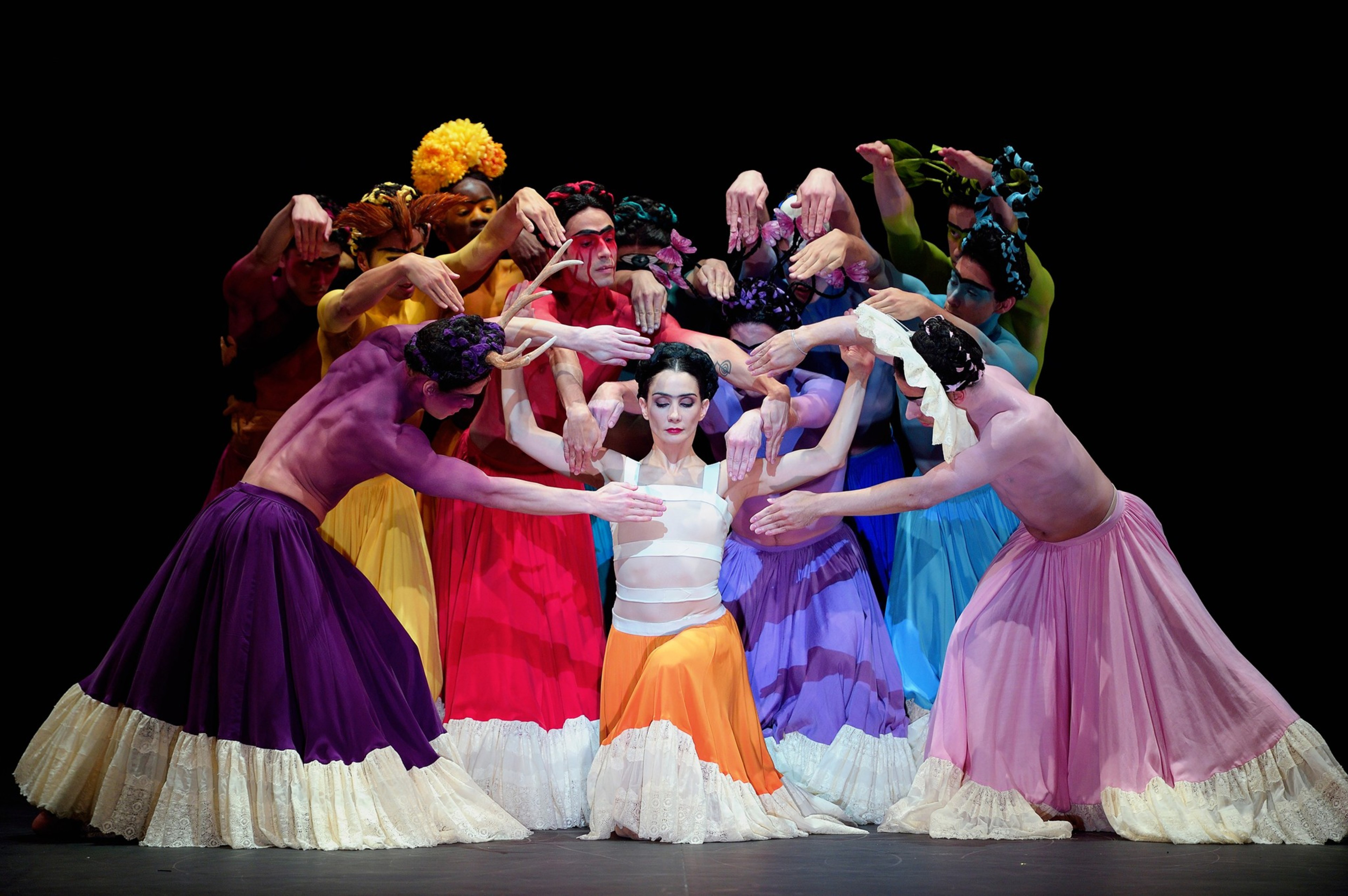 A central dancer in an orange skirt is surrounded by dancers in colorful skirts who extend their arms toward her in a symmetrical, artistic pose against a black background.