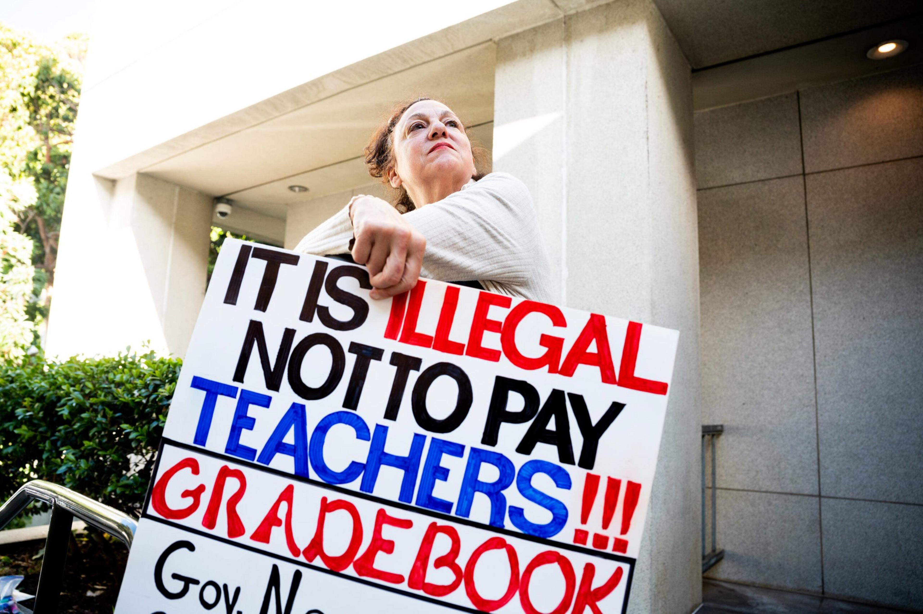 A person holds a sign that says "It is illegal not to pay teachers!!! Gradebook," standing outside a building with a determined expression.