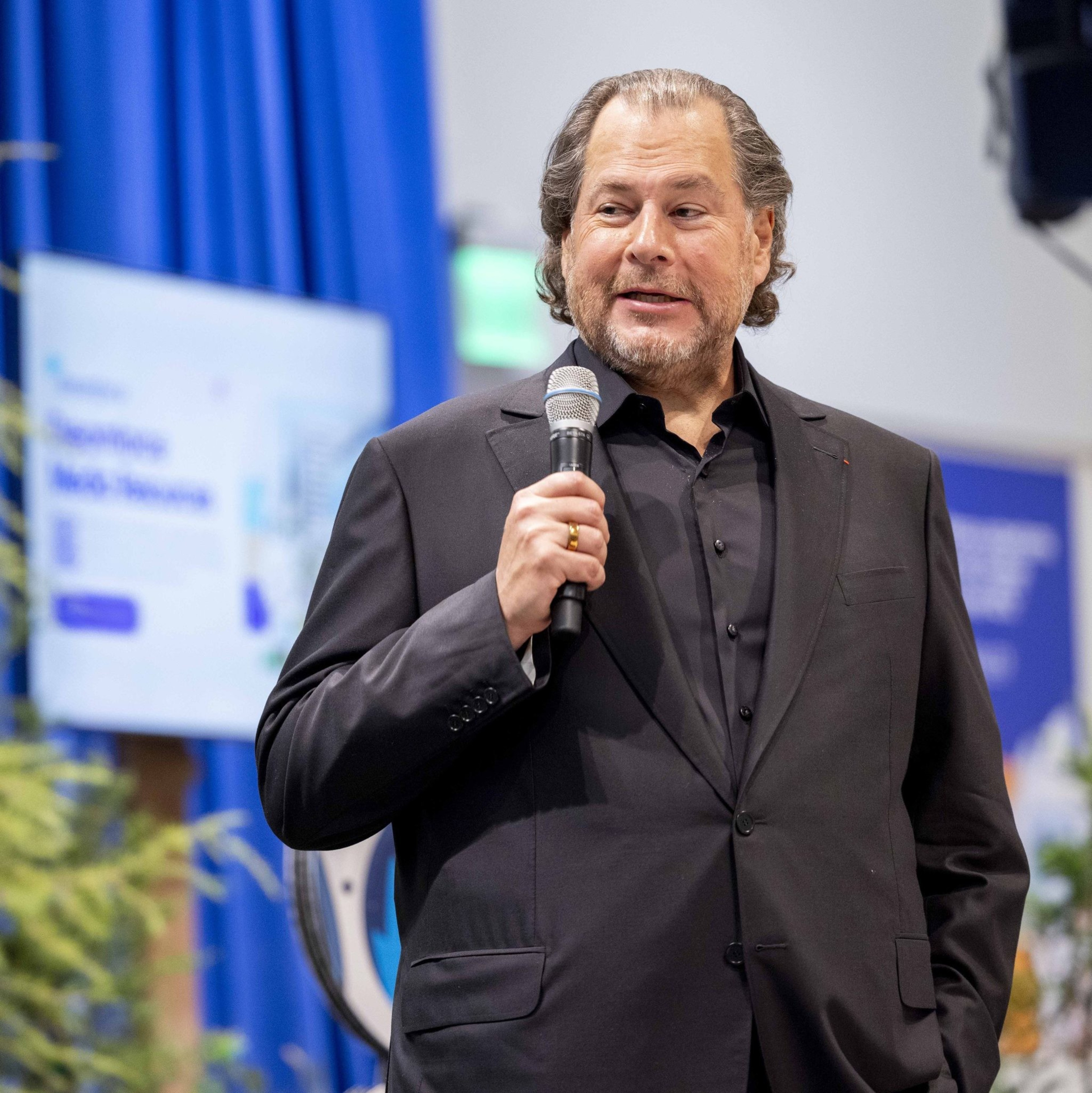A man in a black suit and shirt is speaking into a microphone. He has wavy hair and a beard. The background features blue curtains and a blurred screen.