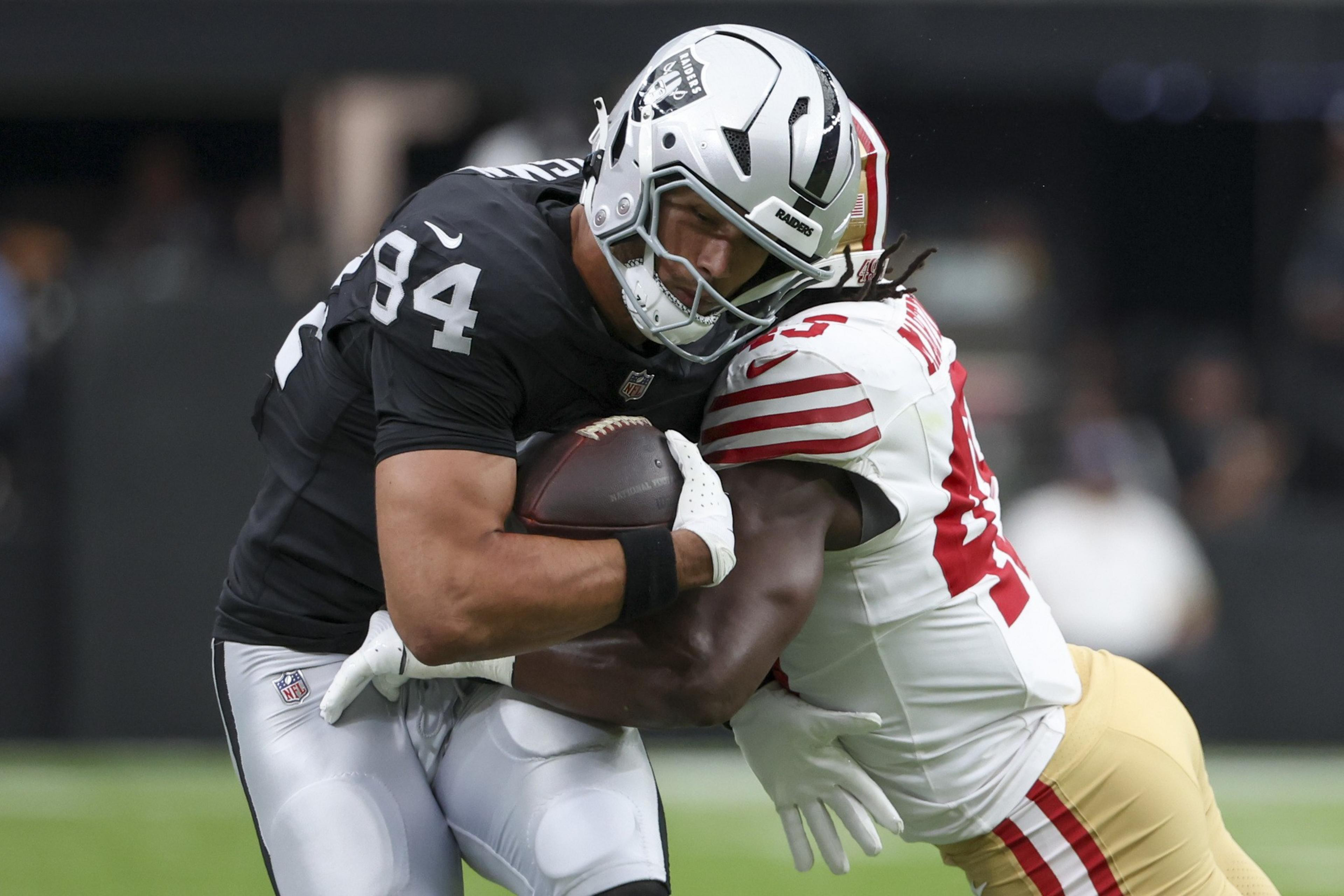 A football player in a black uniform tightly holds the ball while being tackled by an opponent in a white and red uniform.