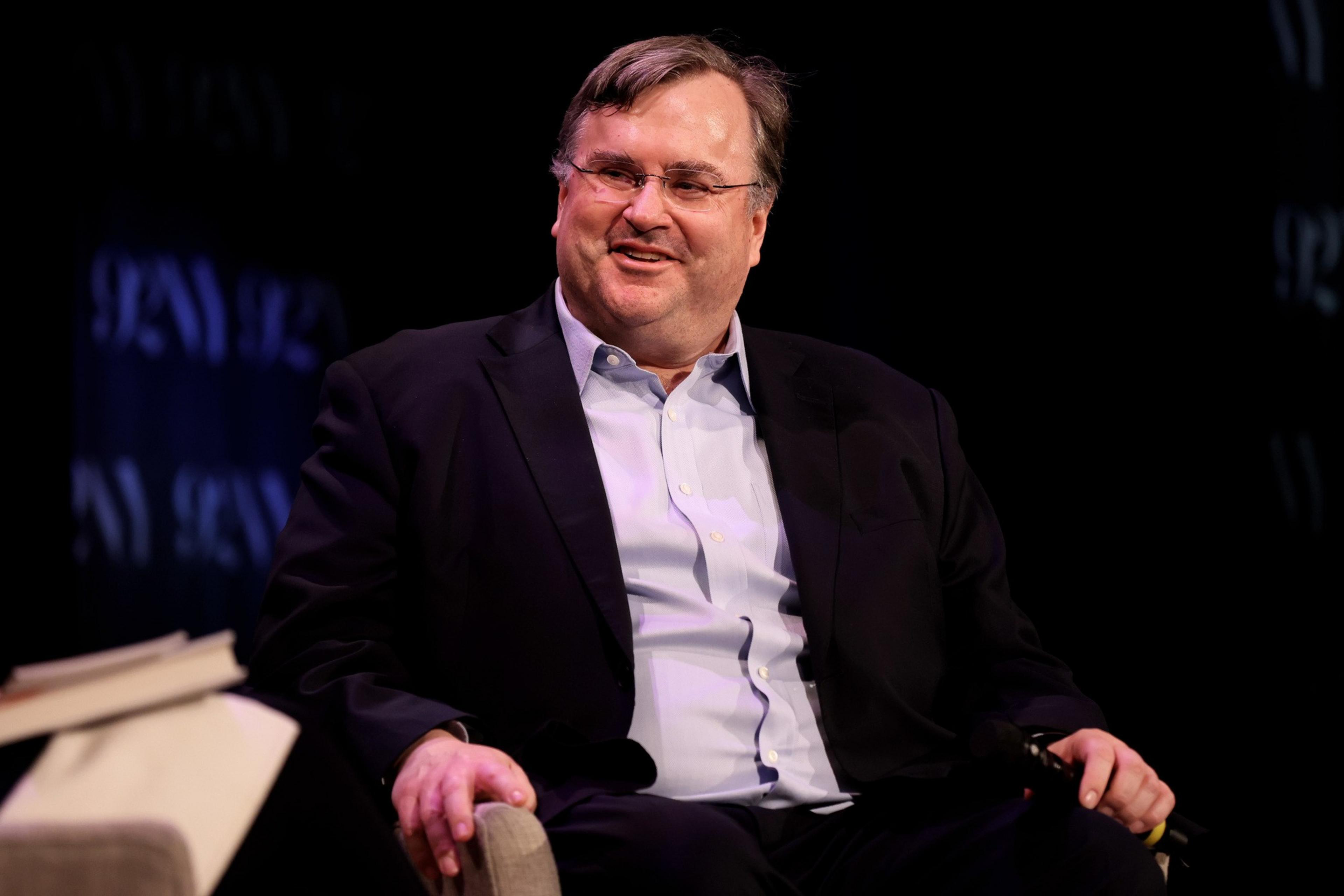 A middle-aged man wearing glasses, a dark blazer, and a light blue shirt sits smiling on a stage with a microphone in hand.