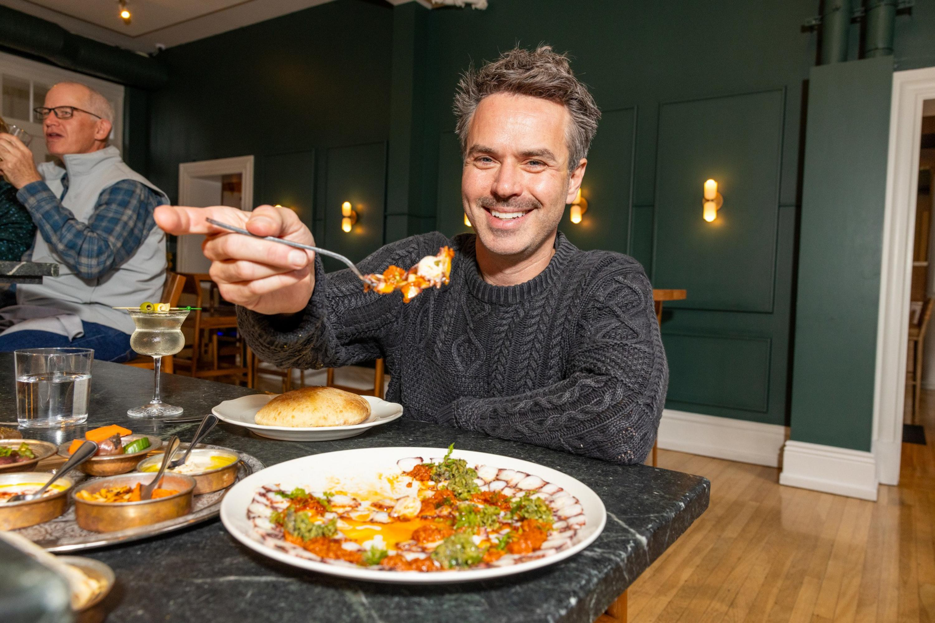 A man wearing a dark sweater smiles while holding a fork with food, sitting at a table with various dishes and a drink in a cozy restaurant.