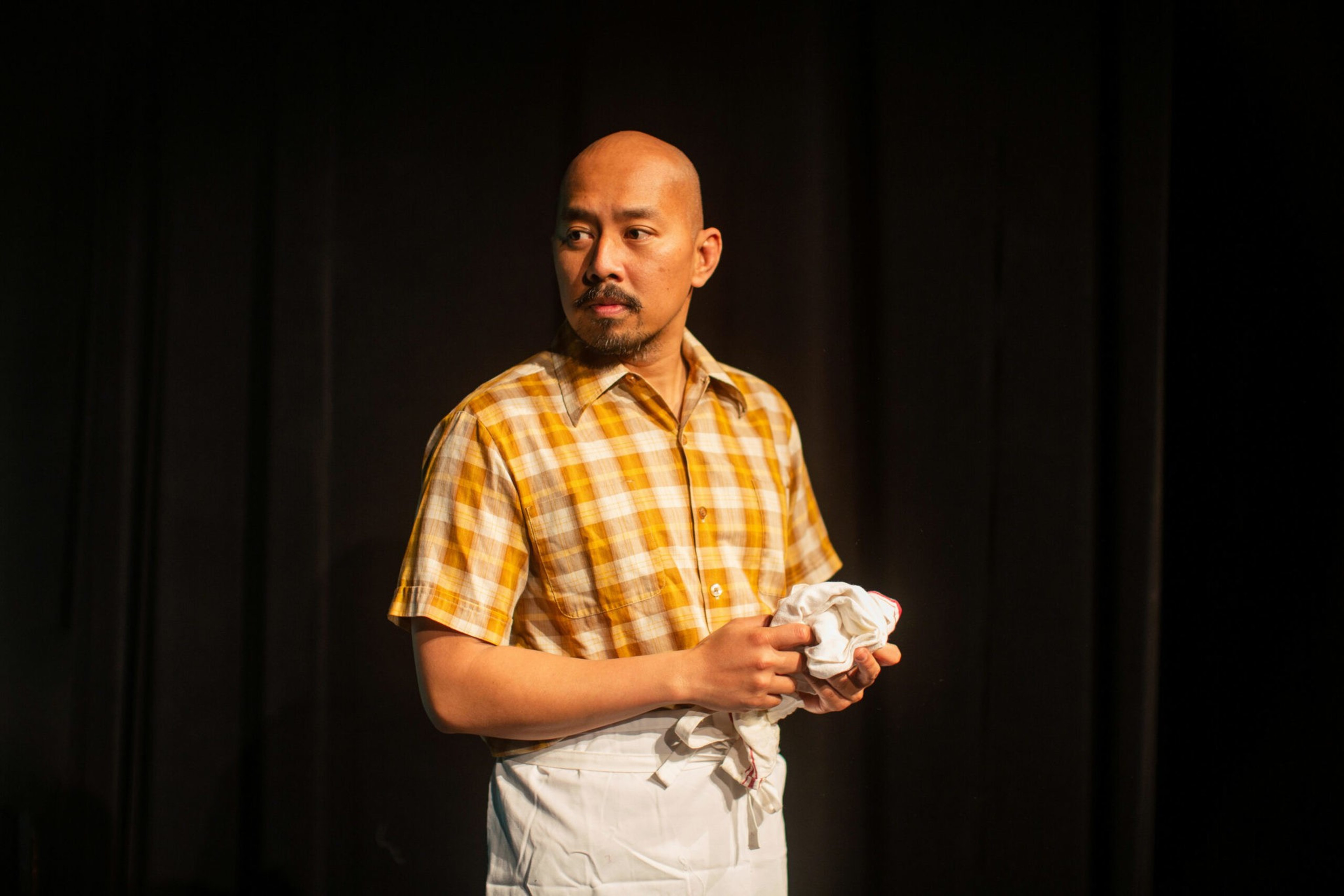 A man in a yellow plaid shirt and white apron holds a cloth, looking thoughtfully to his left against a dark background.