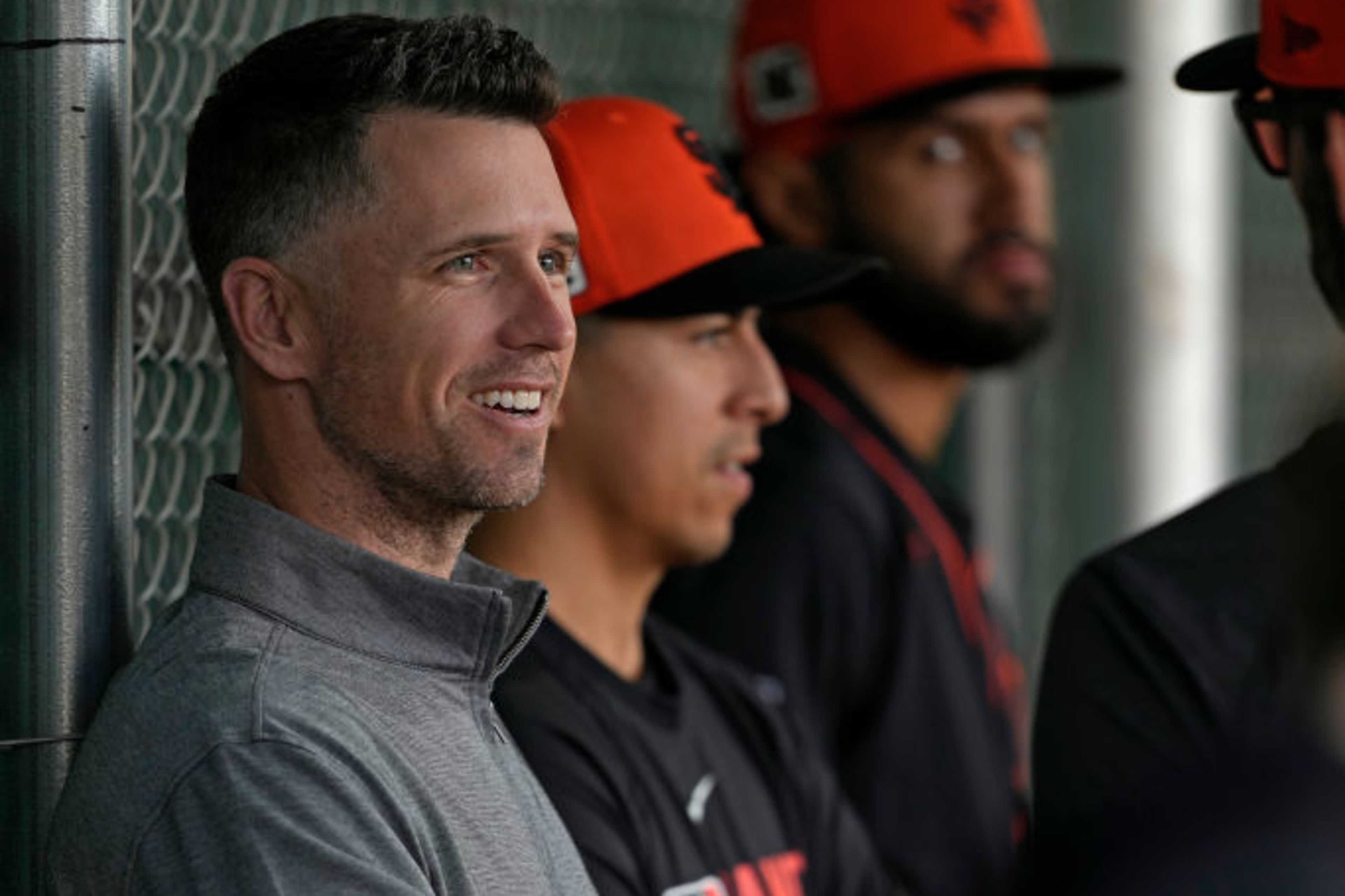 A smiling man in a gray sweater holds a baseball while standing with others in baseball uniforms and orange caps inside a dugout area.