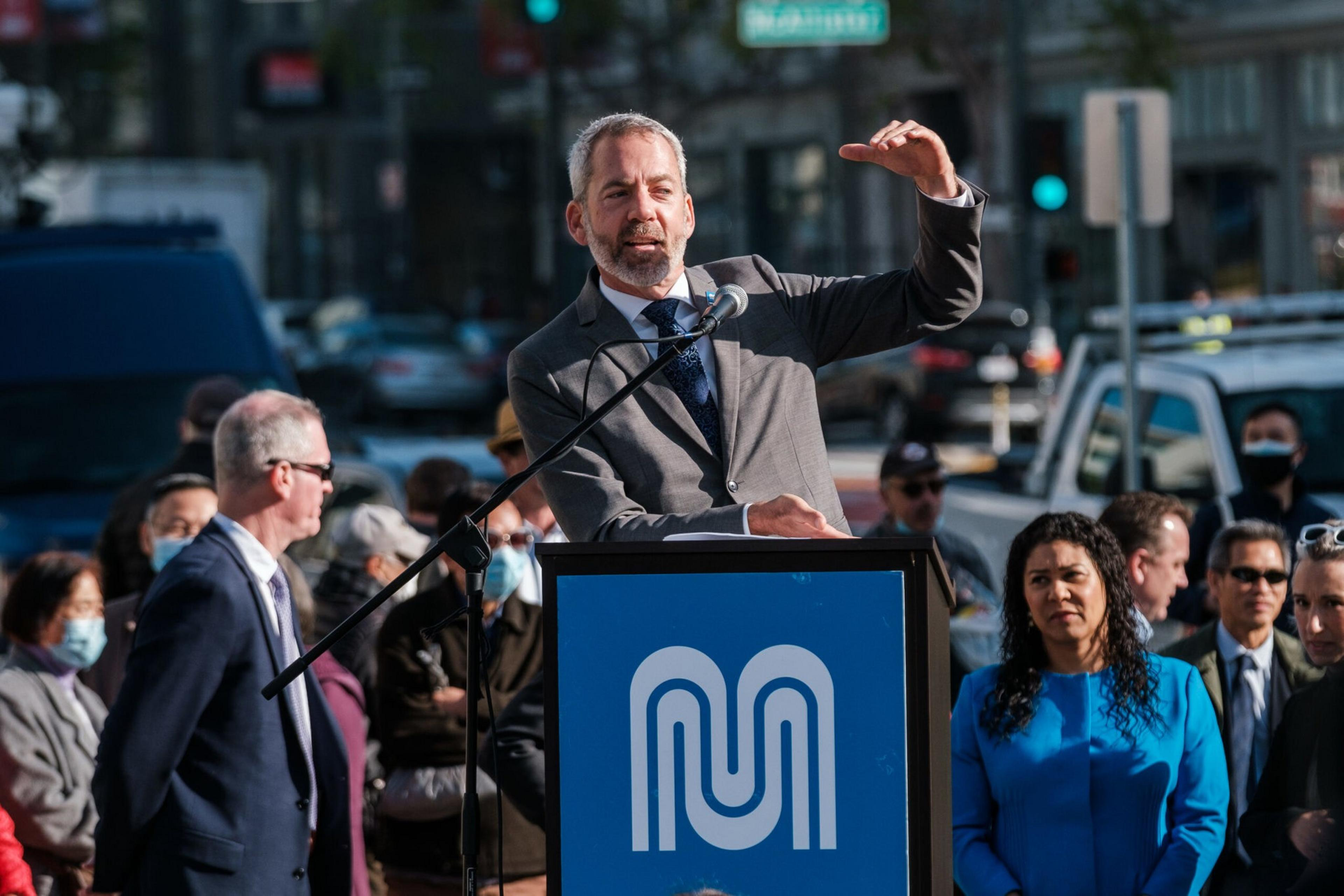 A man speaks at a podium with a blue logo, gesturing upward. A crowd of diverse people, some with masks, listens. Vehicles and a city street are in the background.
