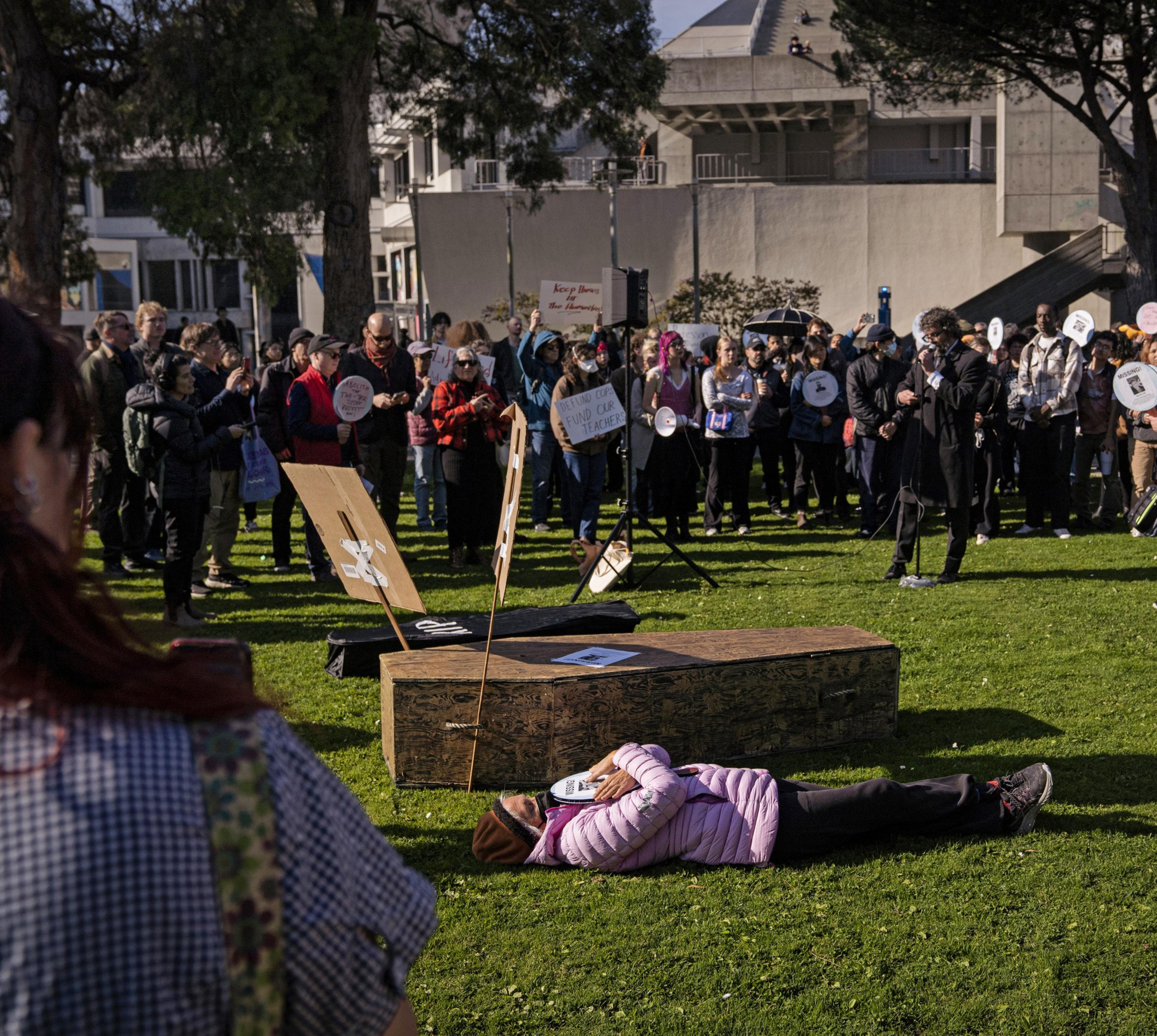 A crowd gathers in a park for a demonstration. A person lies on the grass in front. Signs display messages, and attendees appear engaged and attentive.