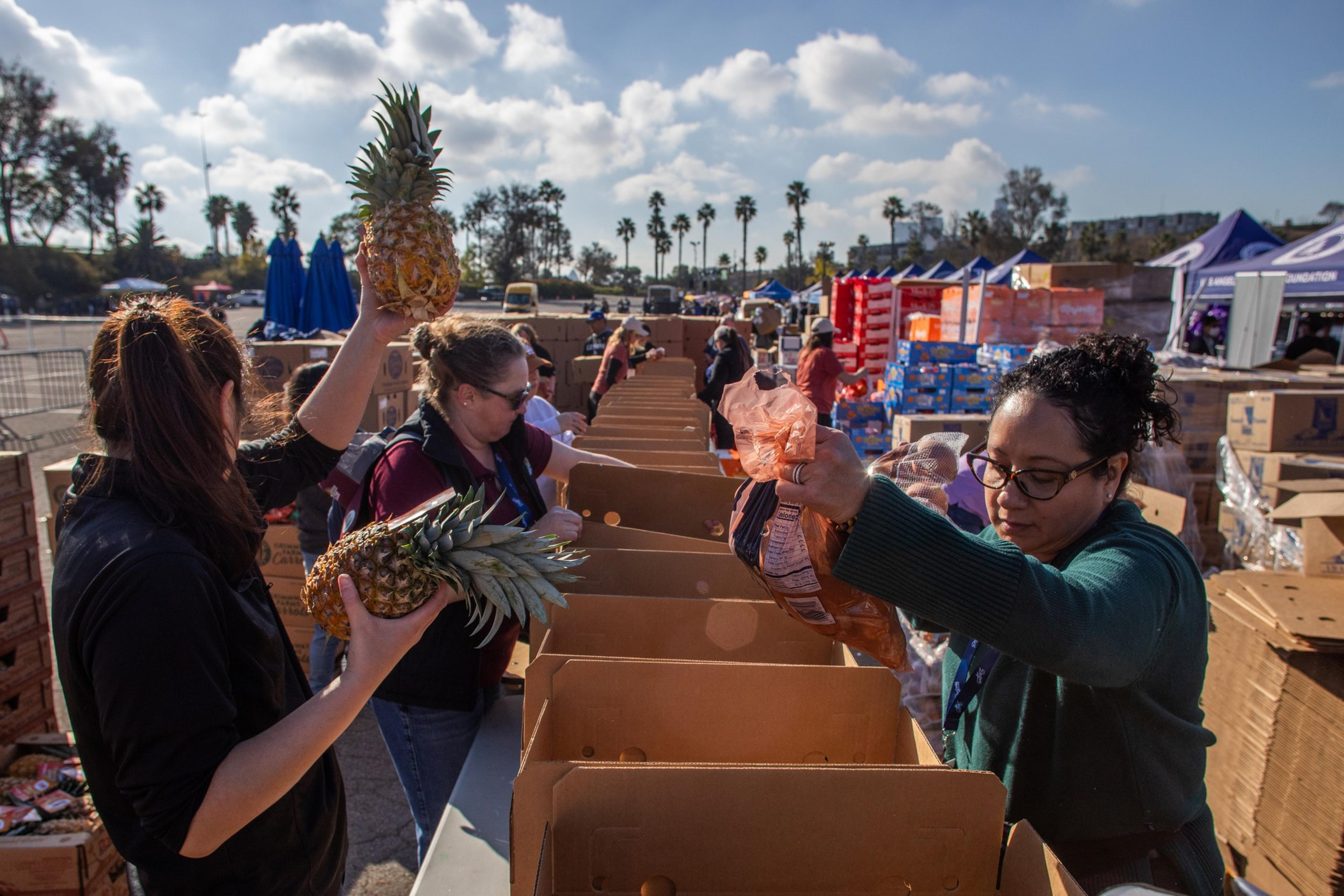 People wearing masks and glasses are packing pineapples and bags of produce into cardboard boxes outdoors under a partly cloudy sky.