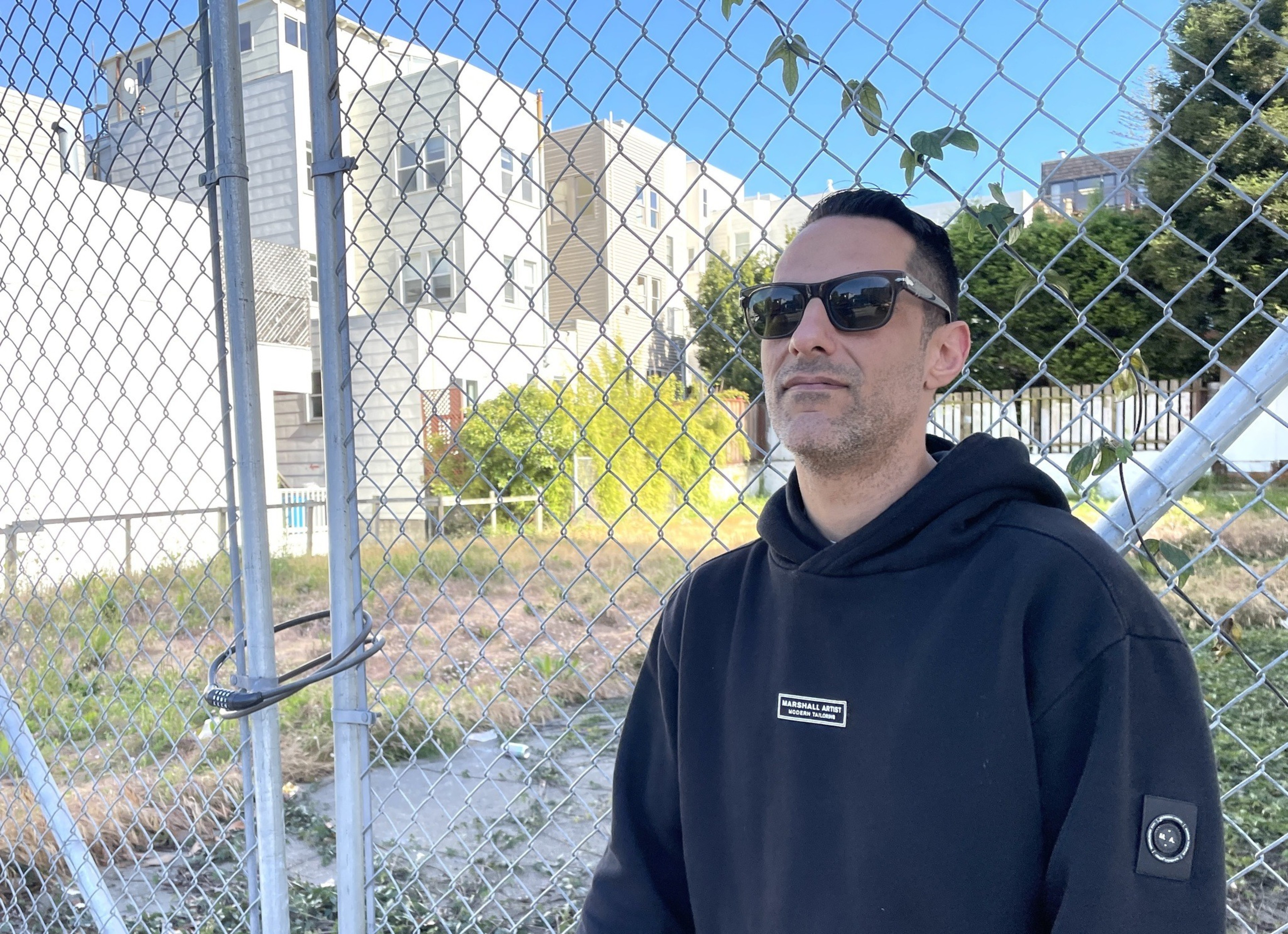 A person wearing sunglasses and a black hoodie stands in front of a chain-link fence. Behind them, there are residential buildings and greenery.