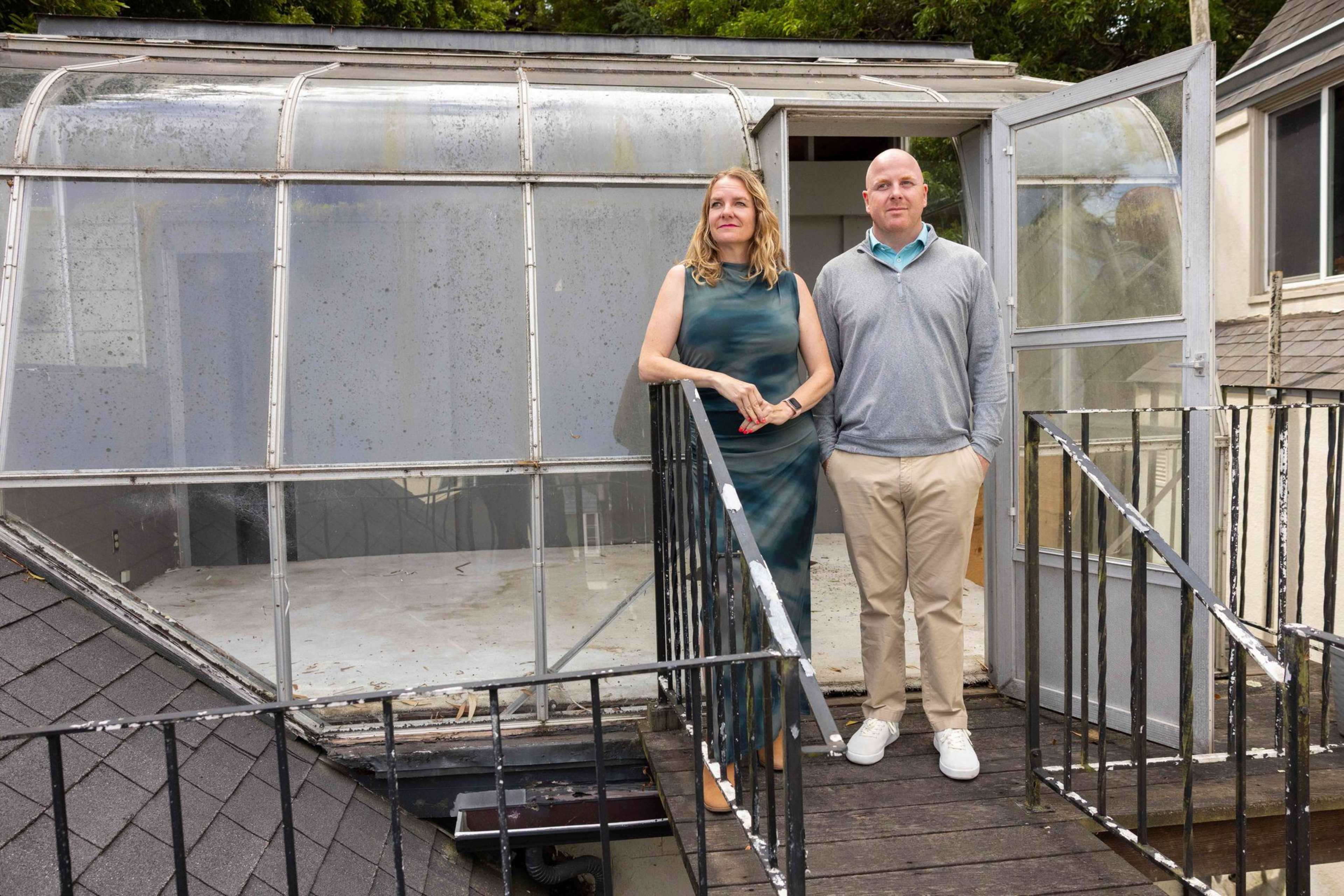 A man and a woman stand side by side on a metal stairway leading to a small, weathered glass enclosure on a rooftop.