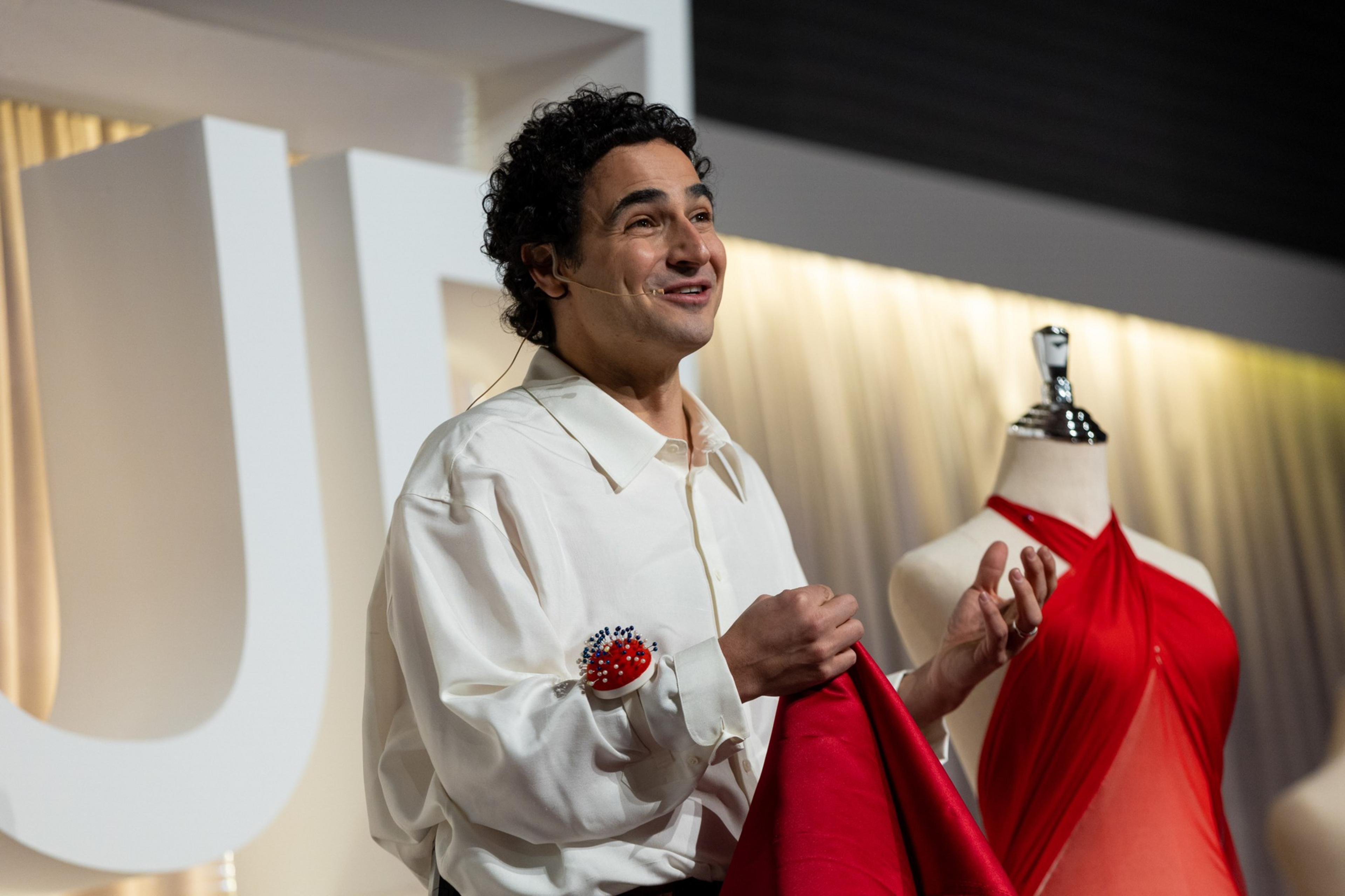 A man in a white shirt gestures near a red dress on a mannequin; large letters and a curtain are in the background.