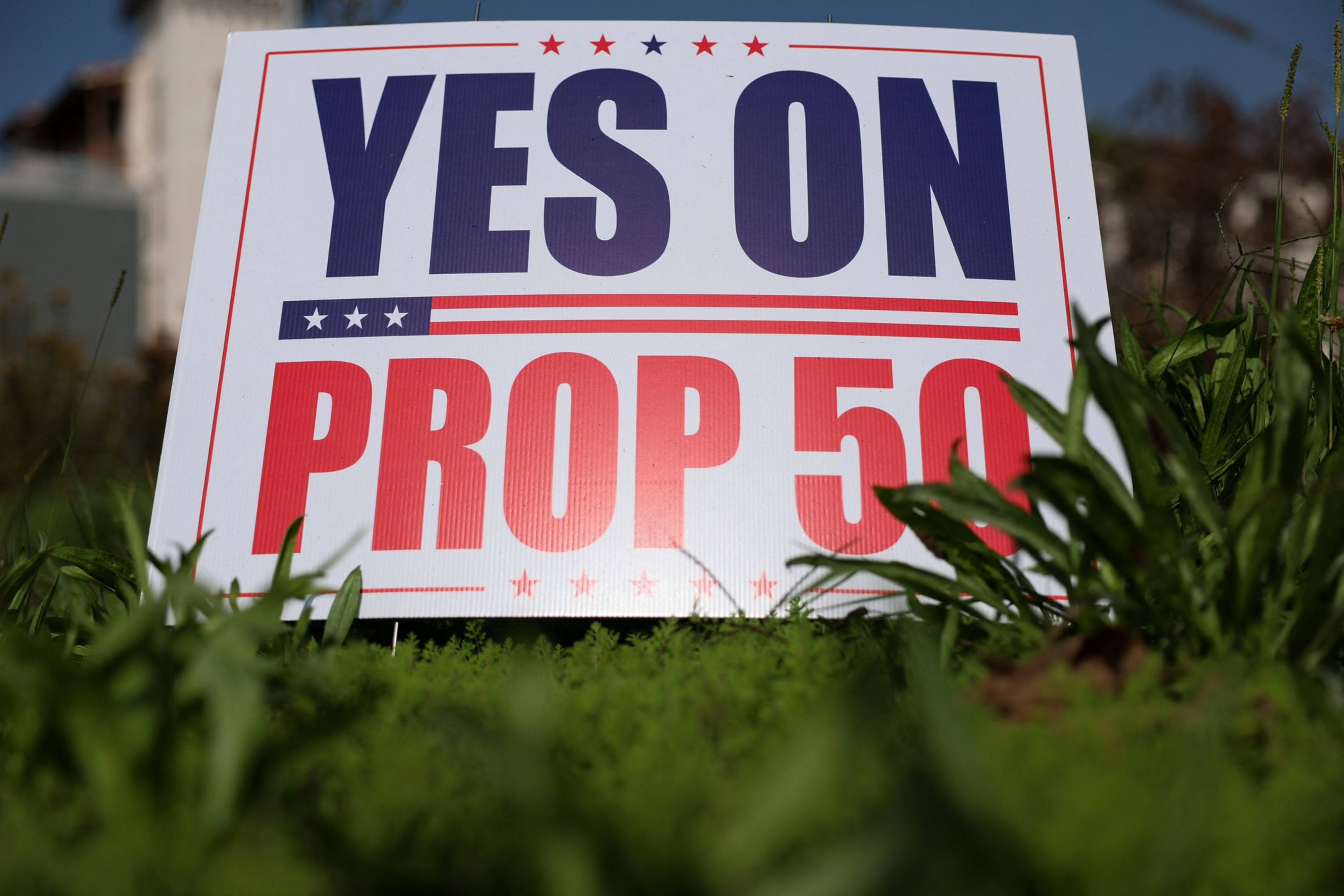 A yard sign with red, white, and blue colors reads "YES ON PROP 50," positioned among green grass and plants.