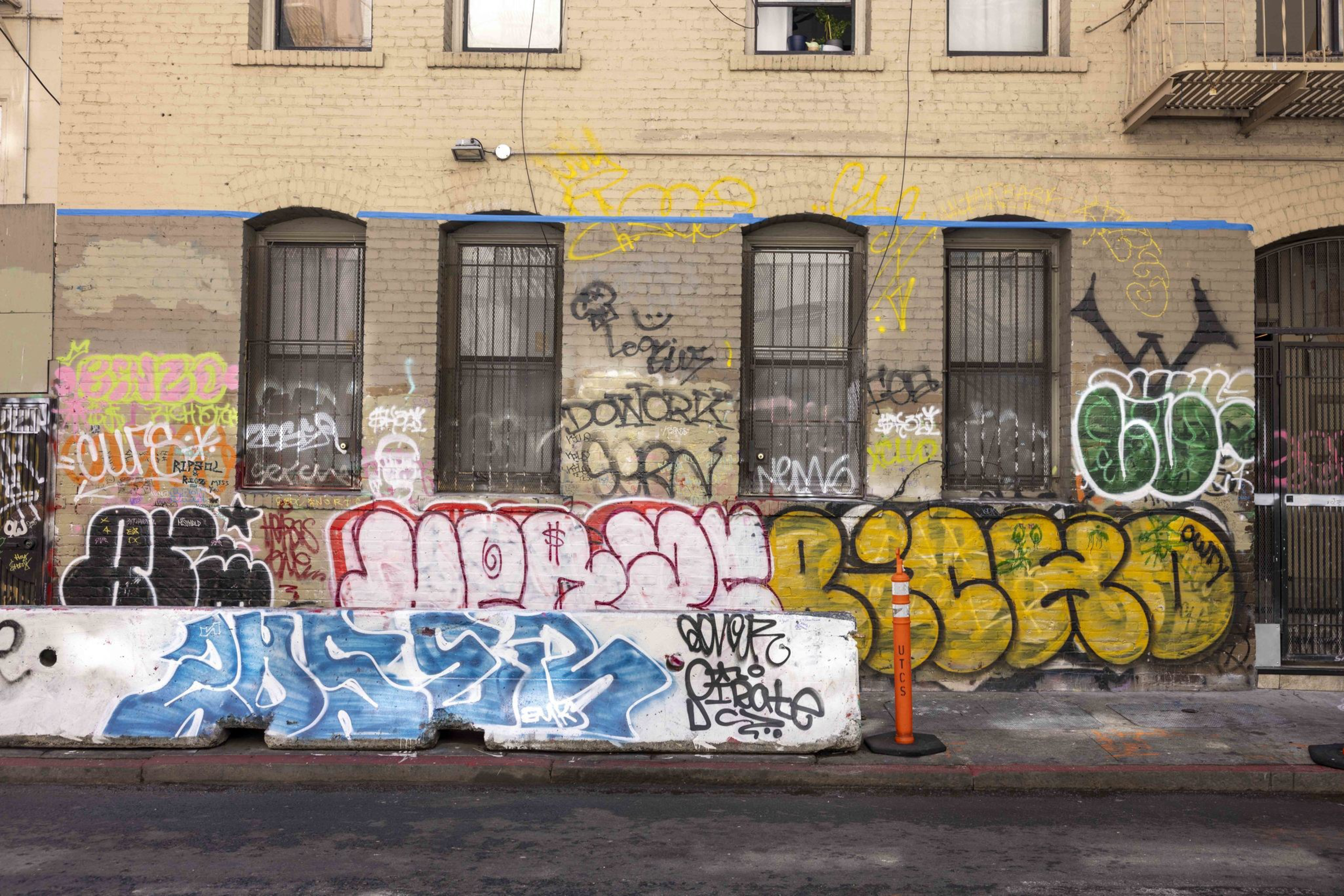The image shows a brick wall with windows, covered in colorful graffiti and tags. A concrete barrier with additional graffiti is in front of the wall.