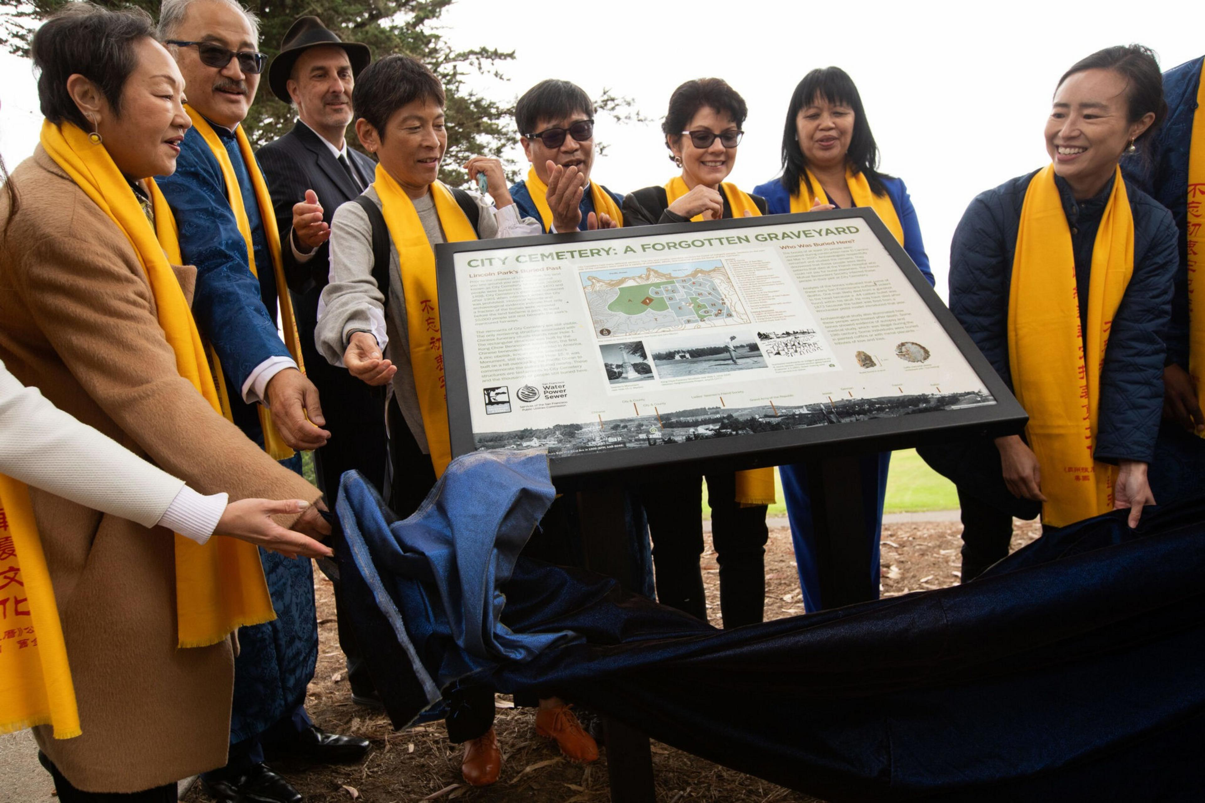 A group of people wearing yellow scarves unveil a historical sign titled "City Cemetery: A Forgotten Graveyard" outdoors.