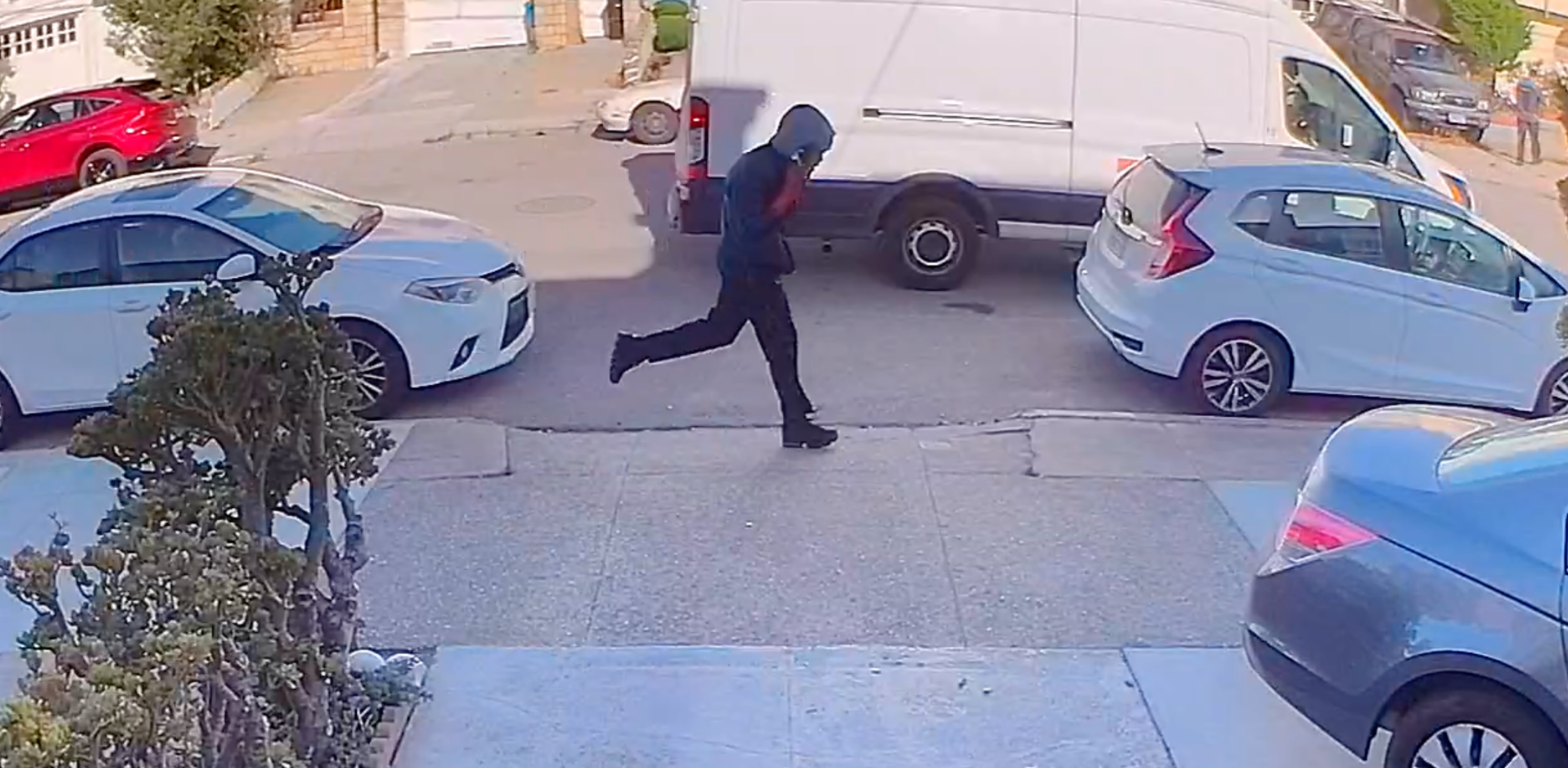 A person wearing a blue hooded jacket and black pants is running across a street lined with parked cars and a white van.