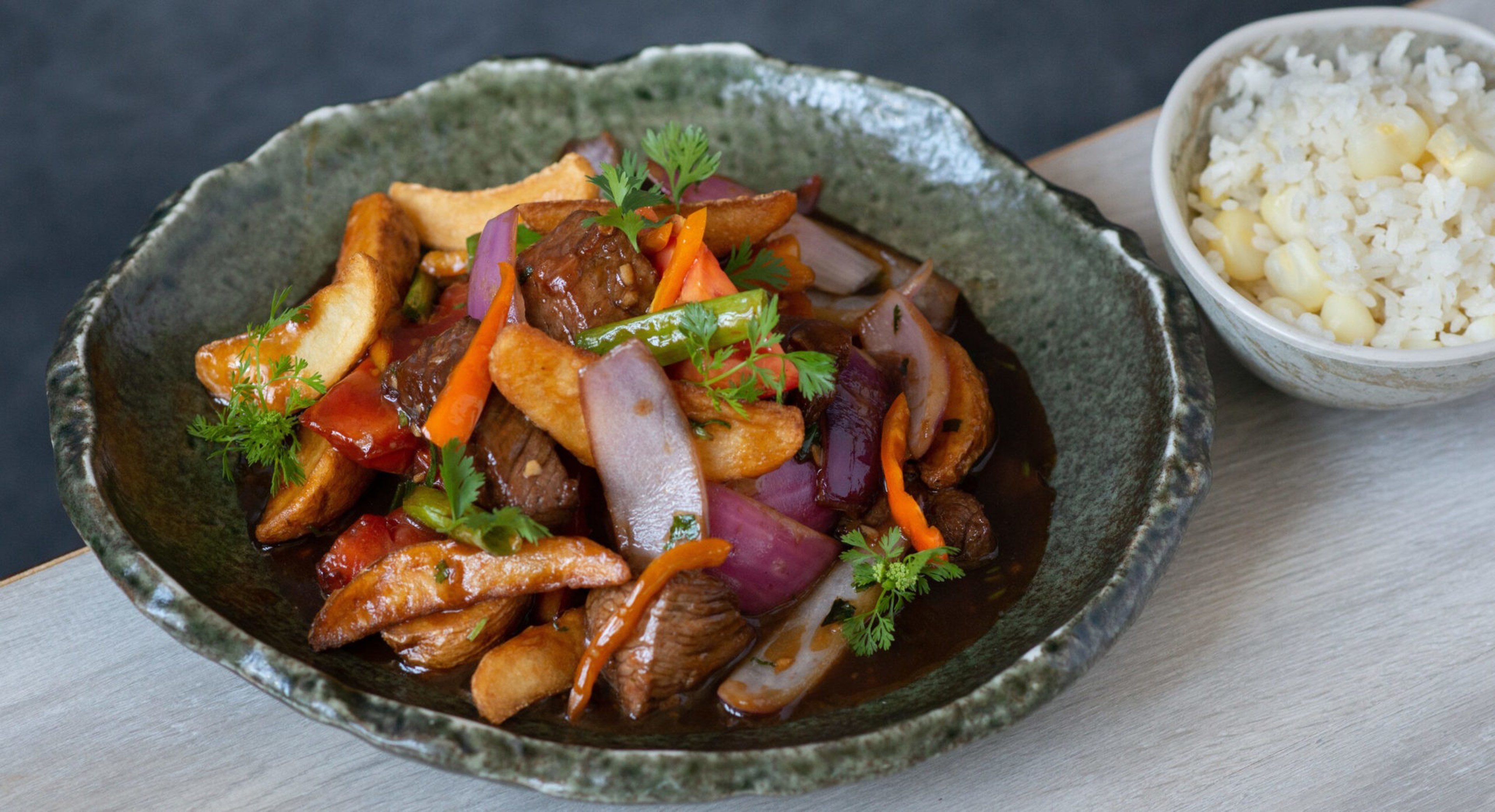 Stir-fried beef with onions, carrots, green peppers, and herbs in a dark sauce, served with a bowl of white rice with corn.